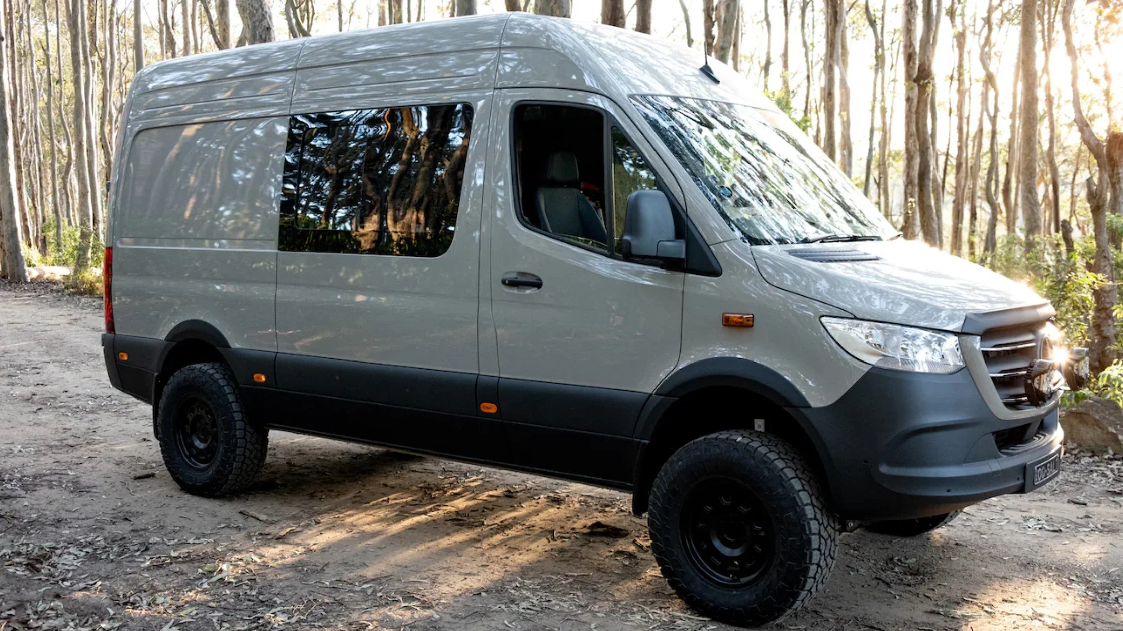 Gray cargo van parked on dirt trail in a woodland area with trees and sunlight