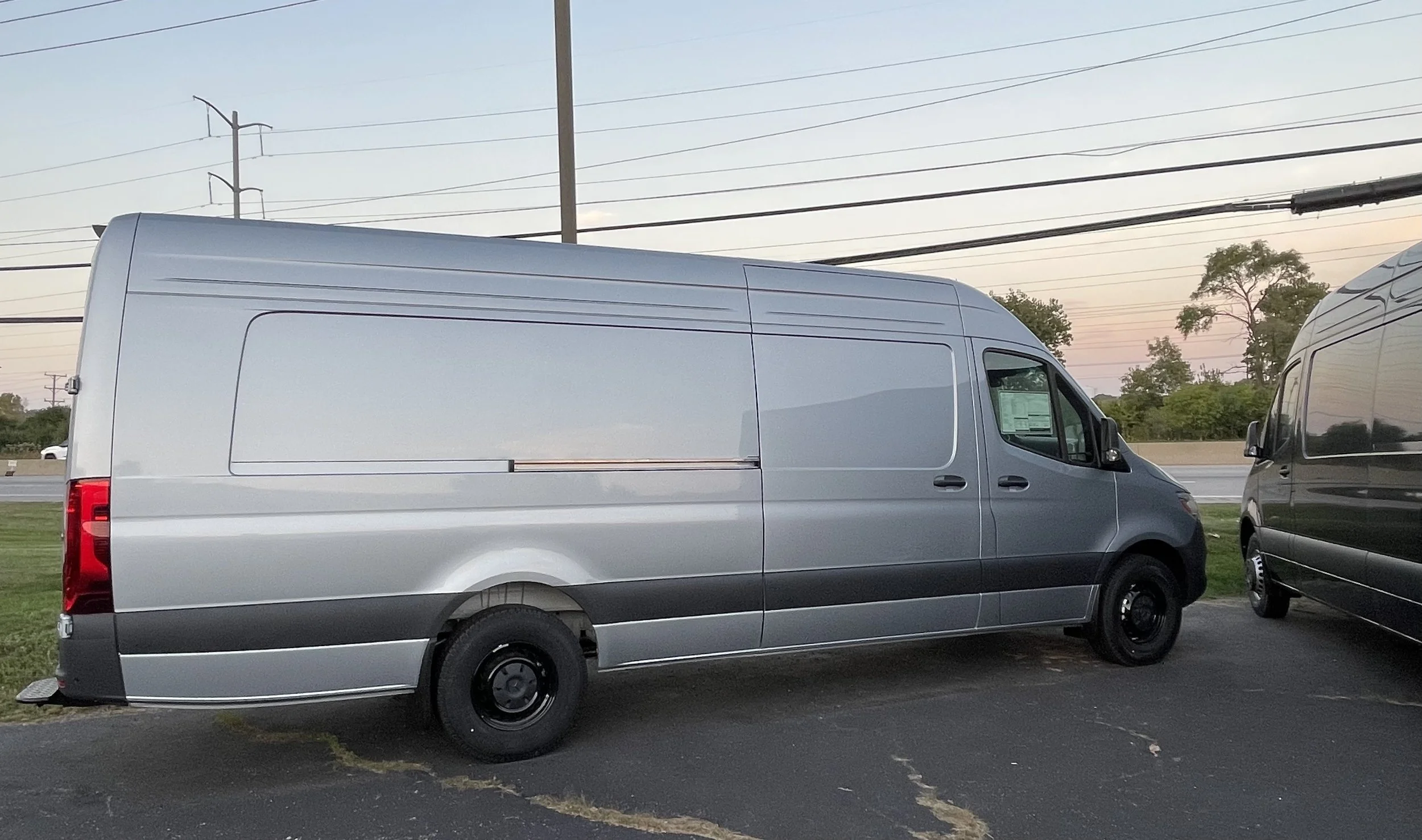 A silver cargo van parked on a paved lot during sunset with trees and power lines in the background.