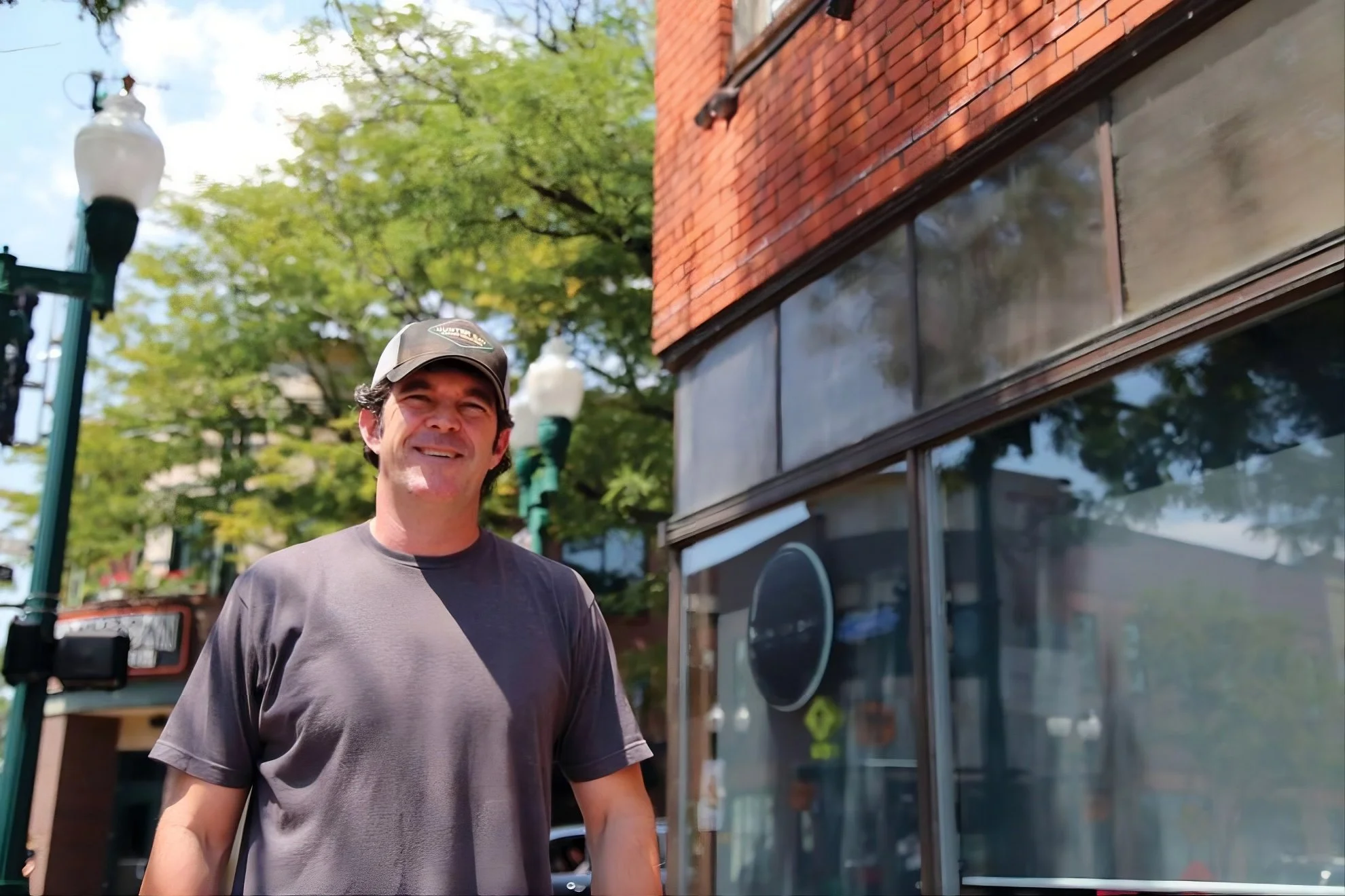 A smiling man in a black cap and gray t-shirt standing outside on a sunny day, with trees, street lamps, and a brick building with large windows reflected in the glass behind him.