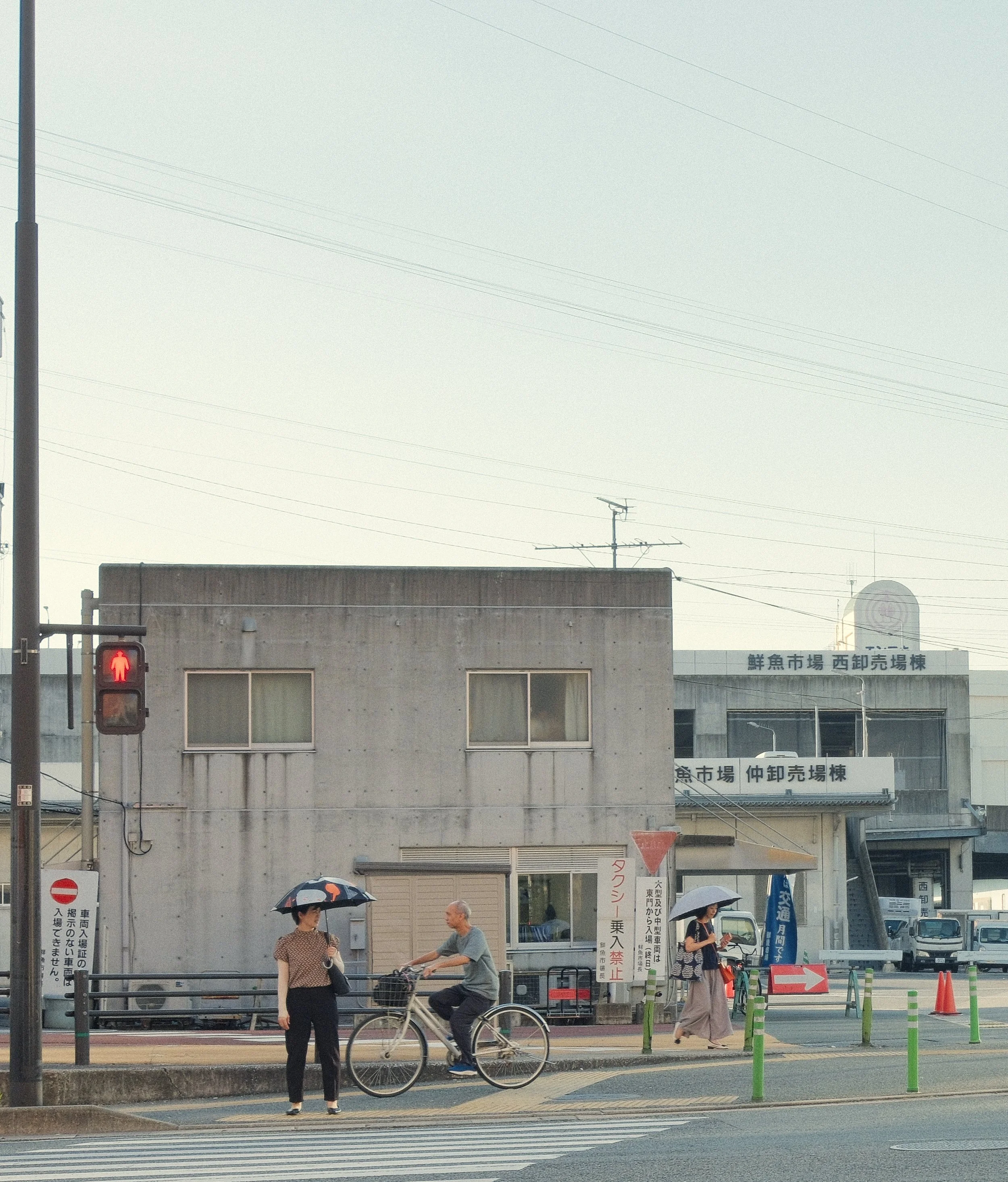 Street scene in Fukuoka, Japan, with pedestrians, a cyclist, traffic lights, and umbrellas on a summer afternoon.