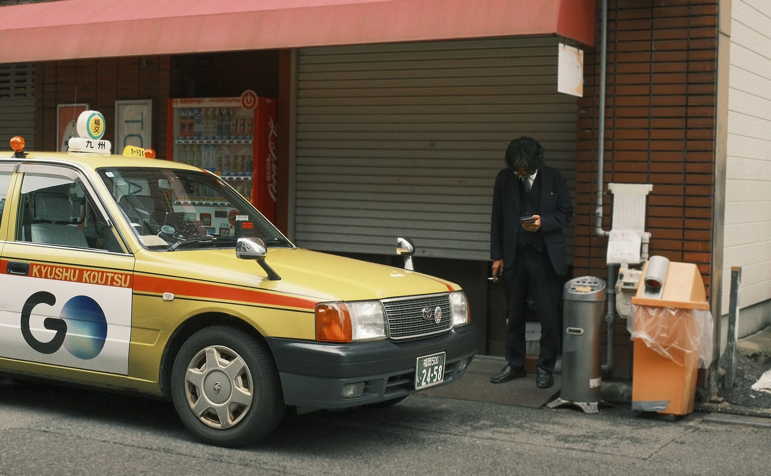Vintage-style taxi on a street in Fukuoka, Japan, with a businessman standing outside a closed storefront.