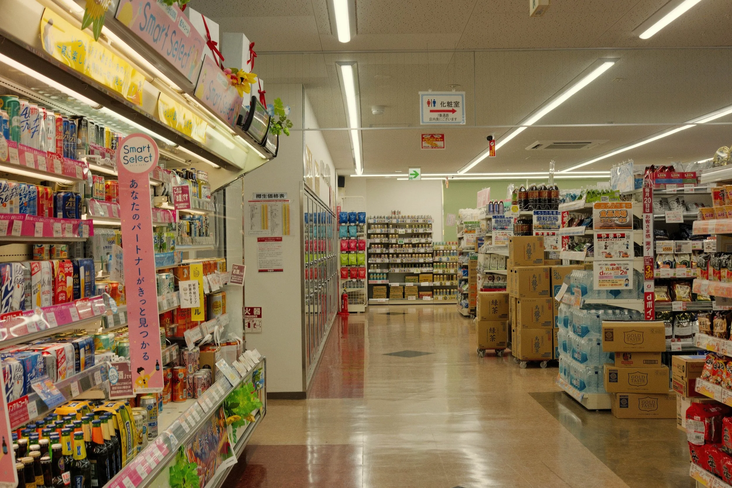 View of grocery store aisle with shelves stocked with drinks, snacks, and household items, featuring signs with Japanese writing, fluorescent lighting, and a clean, organized atmosphere.