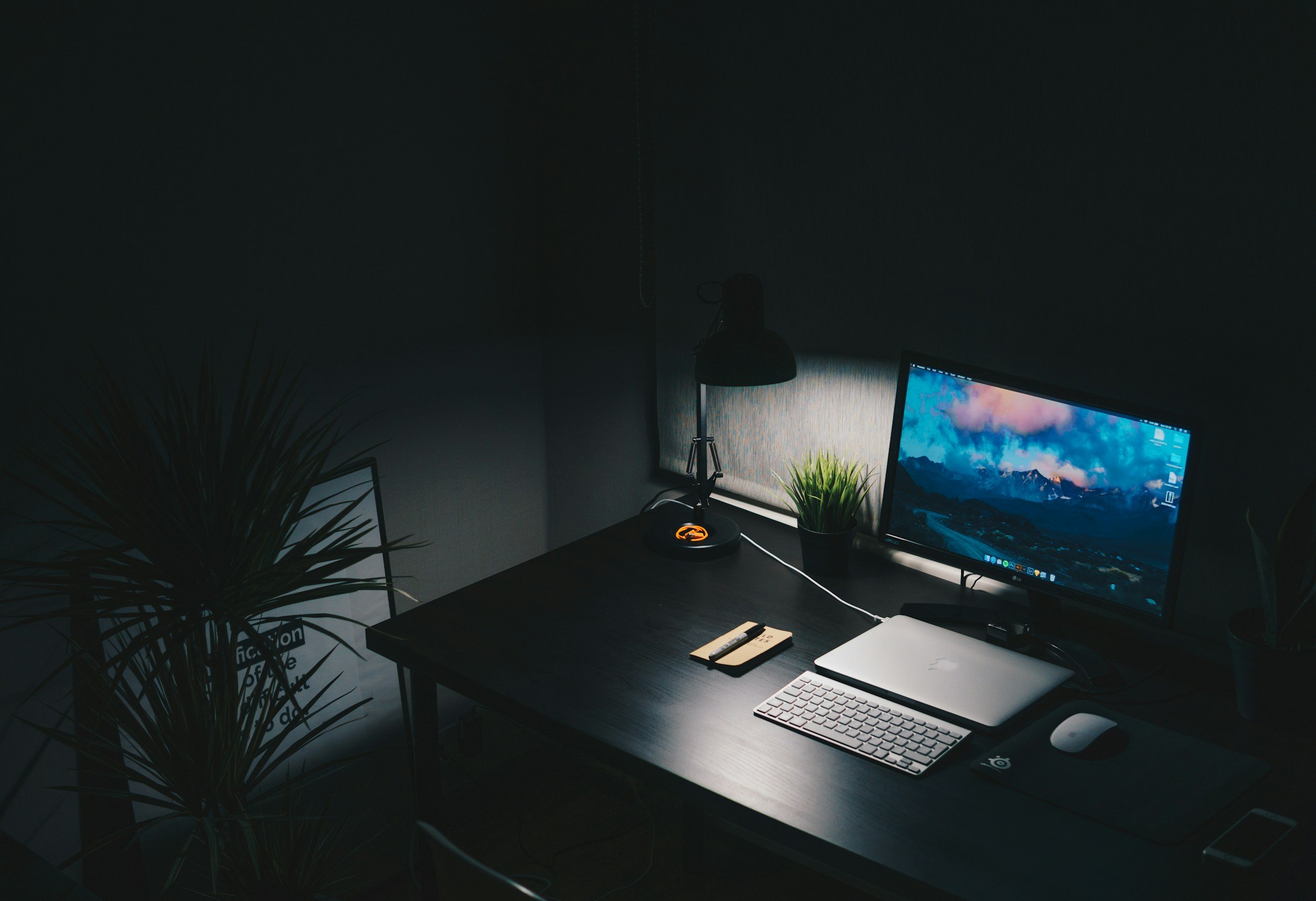 Darkly lit home office with a black desk, a monitor displaying mountain scenery, a keyboard, mouse, closed laptop, desk lamp, small potted plant, notebook with pen, and a framed sign leaning against the wall.