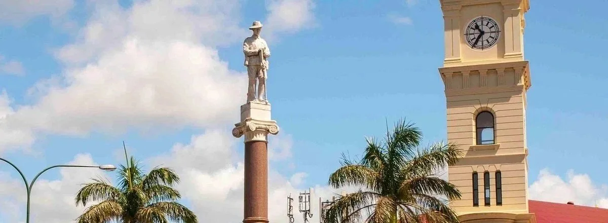 A city square with a tall clock tower, a statue of a historical figure on a column, and palm trees under a partly cloudy sky.