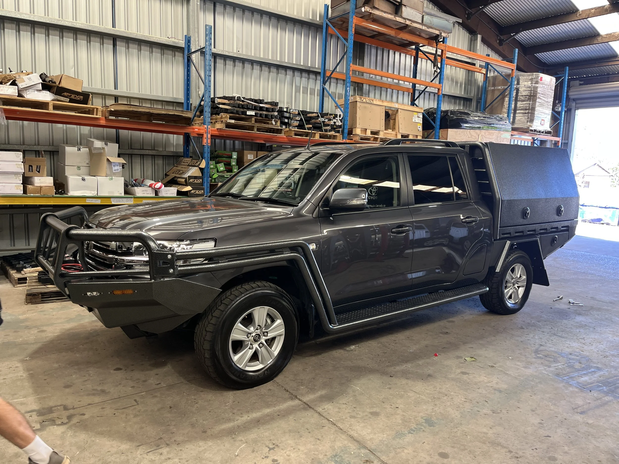A dark gray pickup truck with a custom front bumper and a large black storage box in the bed, parked inside a warehouse with metal shelving and cardboard boxes.