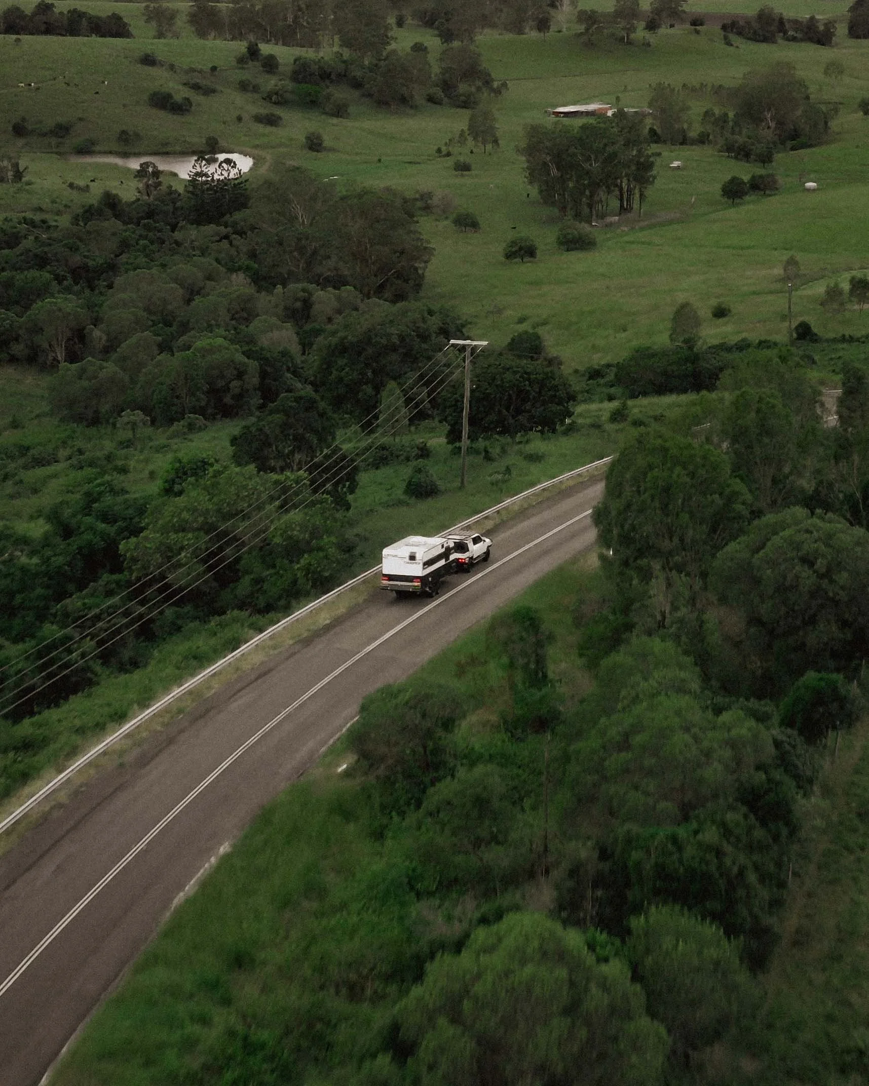 A white truck towing a camper traveling along a winding rural road surrounded by lush green trees and open fields.