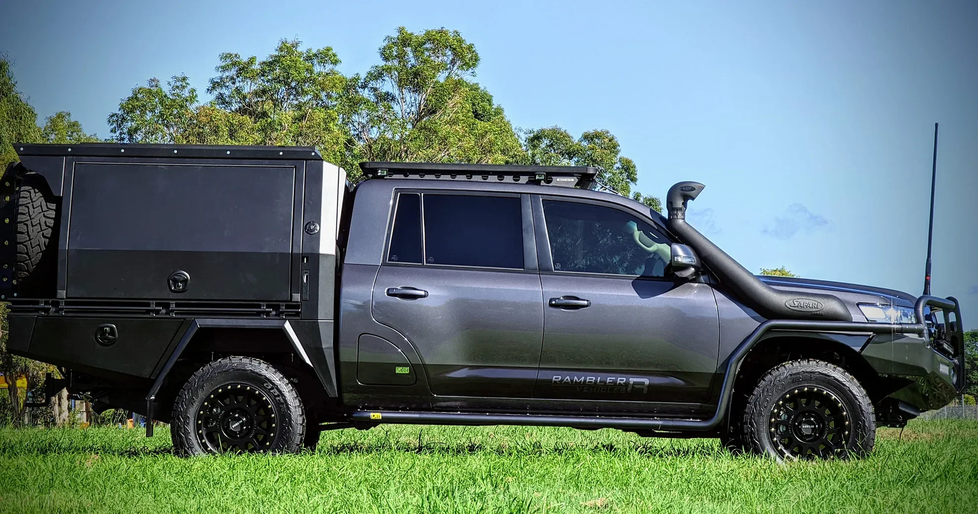 Black off-road utility vehicle with cargo storage box on back, large tires, snorkel, and side steps, parked on grassy field under blue sky with trees in background.