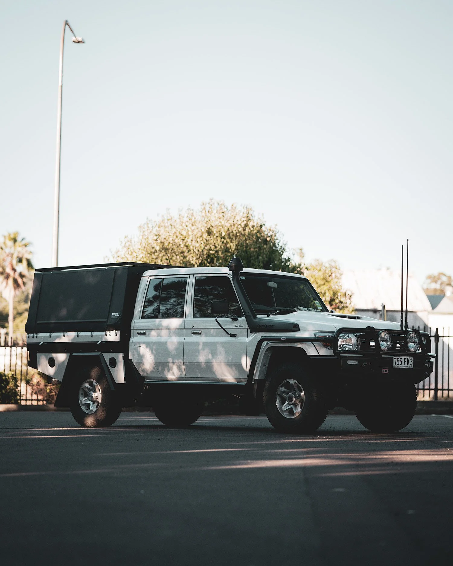 A white four-door off-road vehicle with chrome wheels parked on a paved lot under a clear sky, with a black cargo cover on the back and two tall antennae on the front bumper.