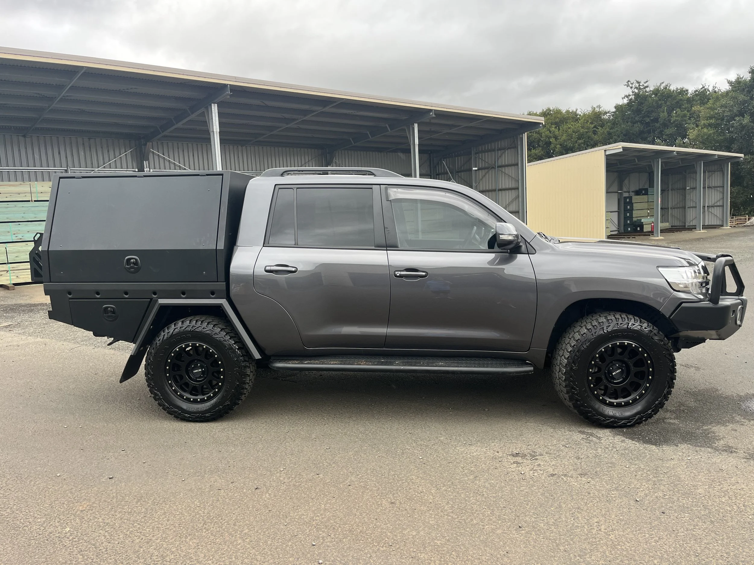 Gray pickup truck with a custom cargo box, off-road tires, and a front bull bar parked on a paved surface with metal storage sheds in the background under a cloudy sky.