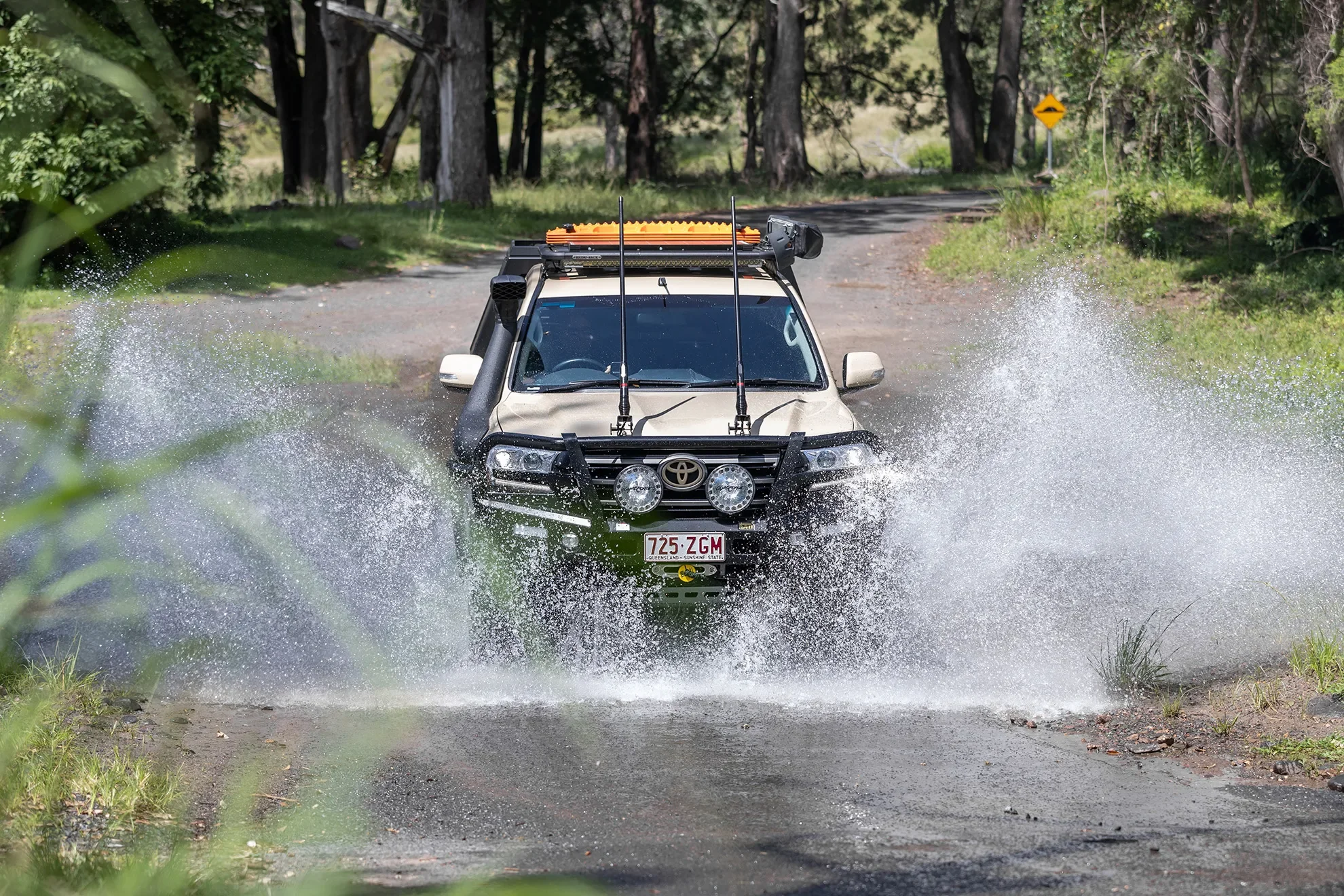 Off-road vehicle driving through a water crossing on a dirt trail surrounded by trees.