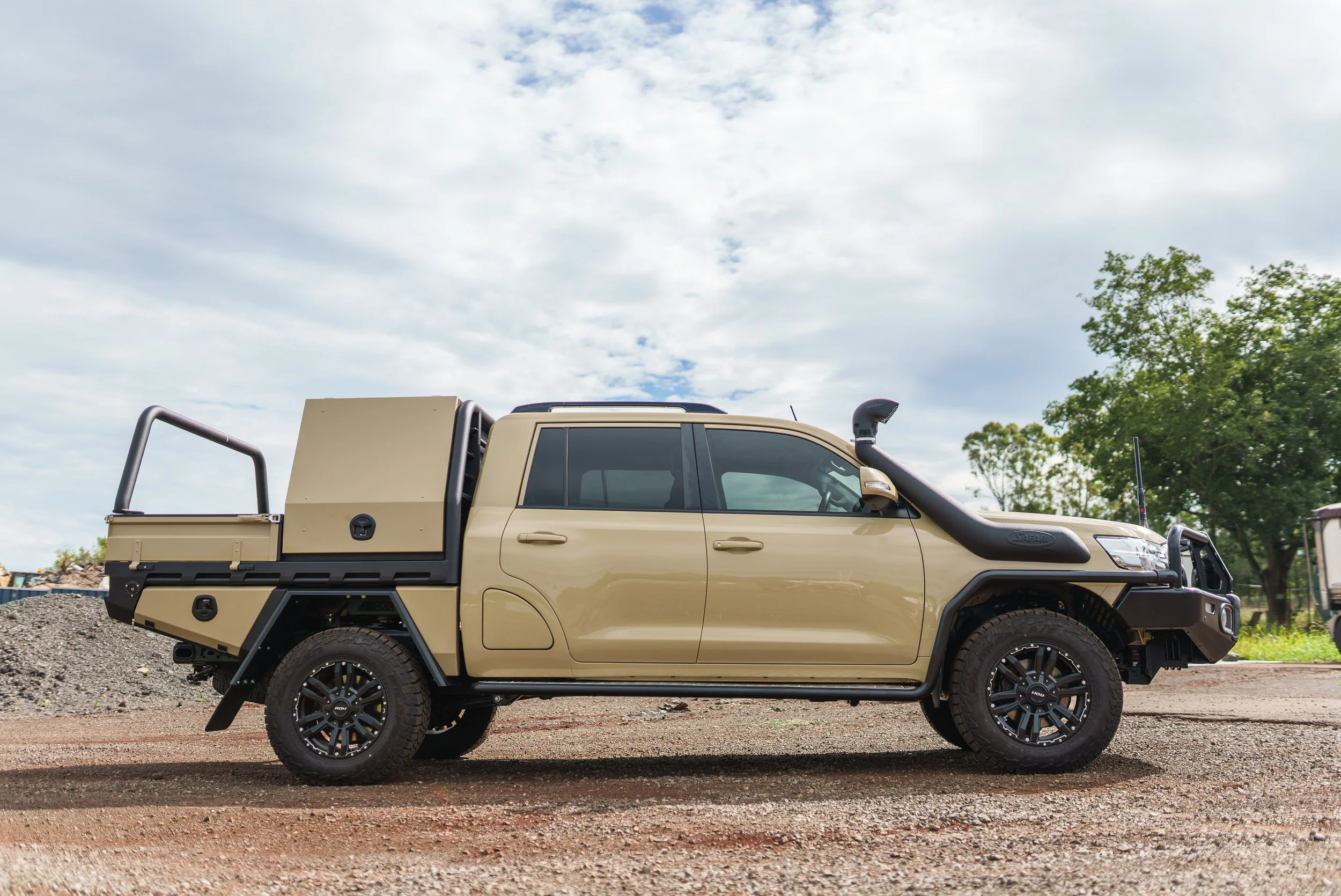 A beige pickup truck with a black off-road bumper, snorkel, and rugged tires parked on a gravel lot under a partly cloudy sky, with trees in the background.