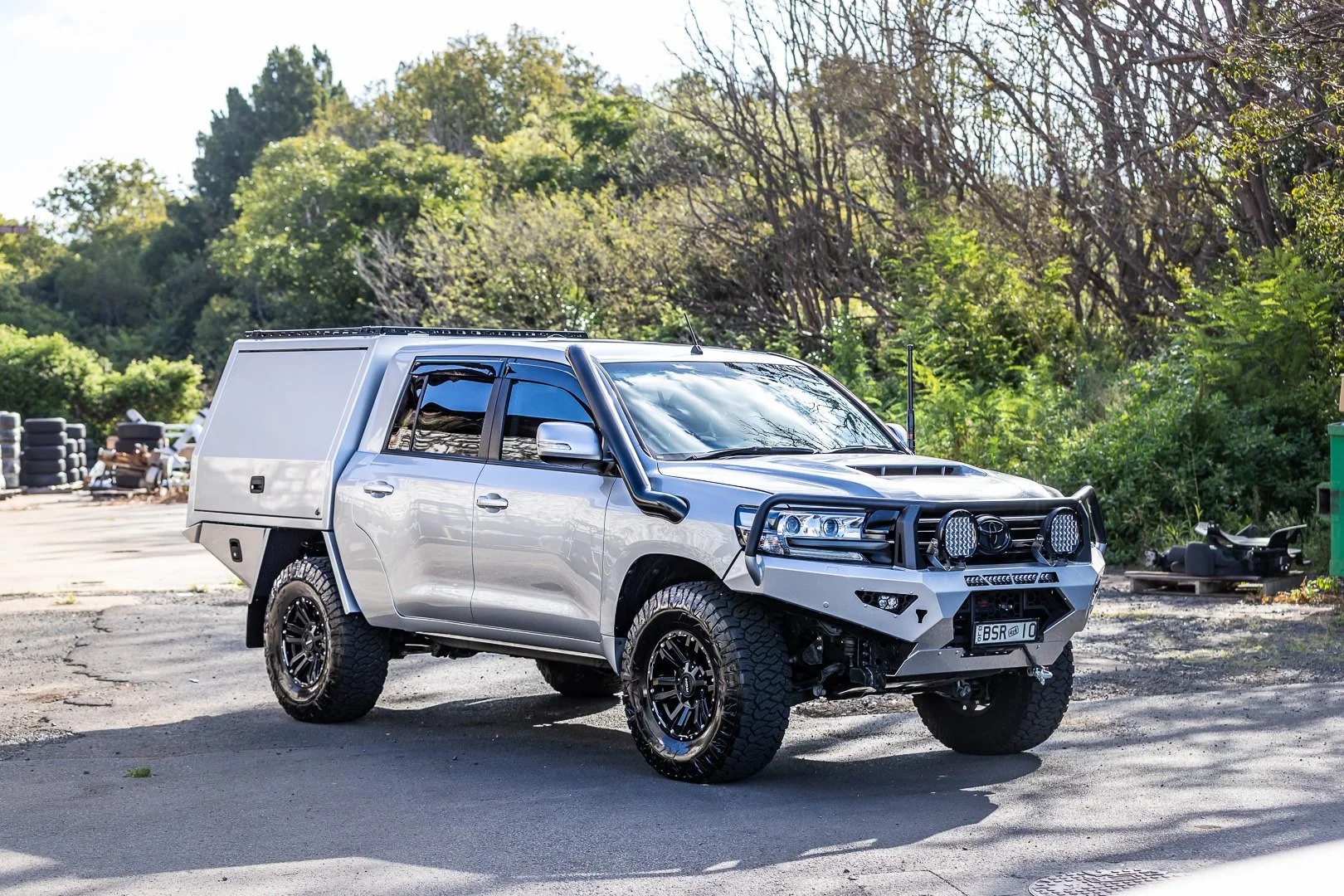 Silver off-road utility vehicle with a canopy, large tires, and front bull bars parked on a paved surface with greenery and trees in the background.