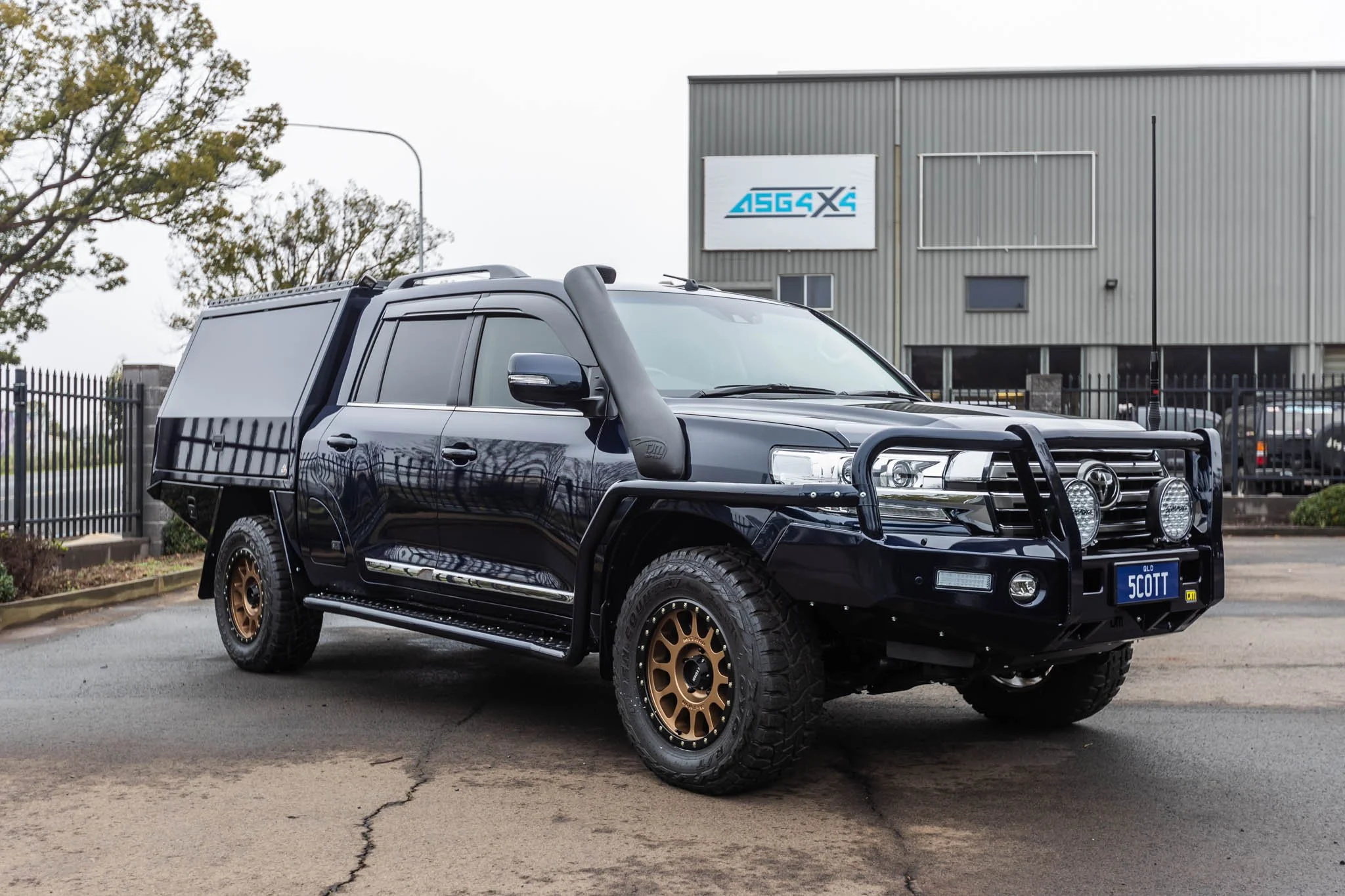 Black pickup truck with off-road tires, bull bar, and canopy parked in front of a gray industrial building with a sign reading AEGAS.