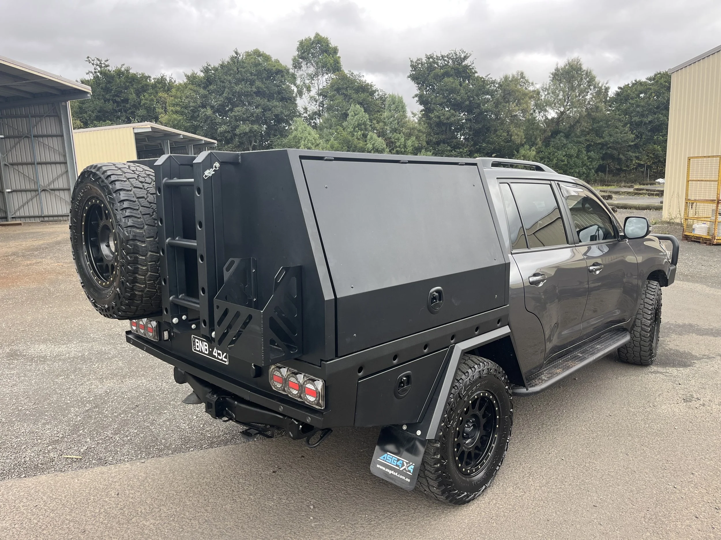Black SUV with a large black cargo box attached to the back, spare tire mounted on the cargo box, off-road tires, and side steps, parked on an outdoor lot with industrial buildings and trees in the background.