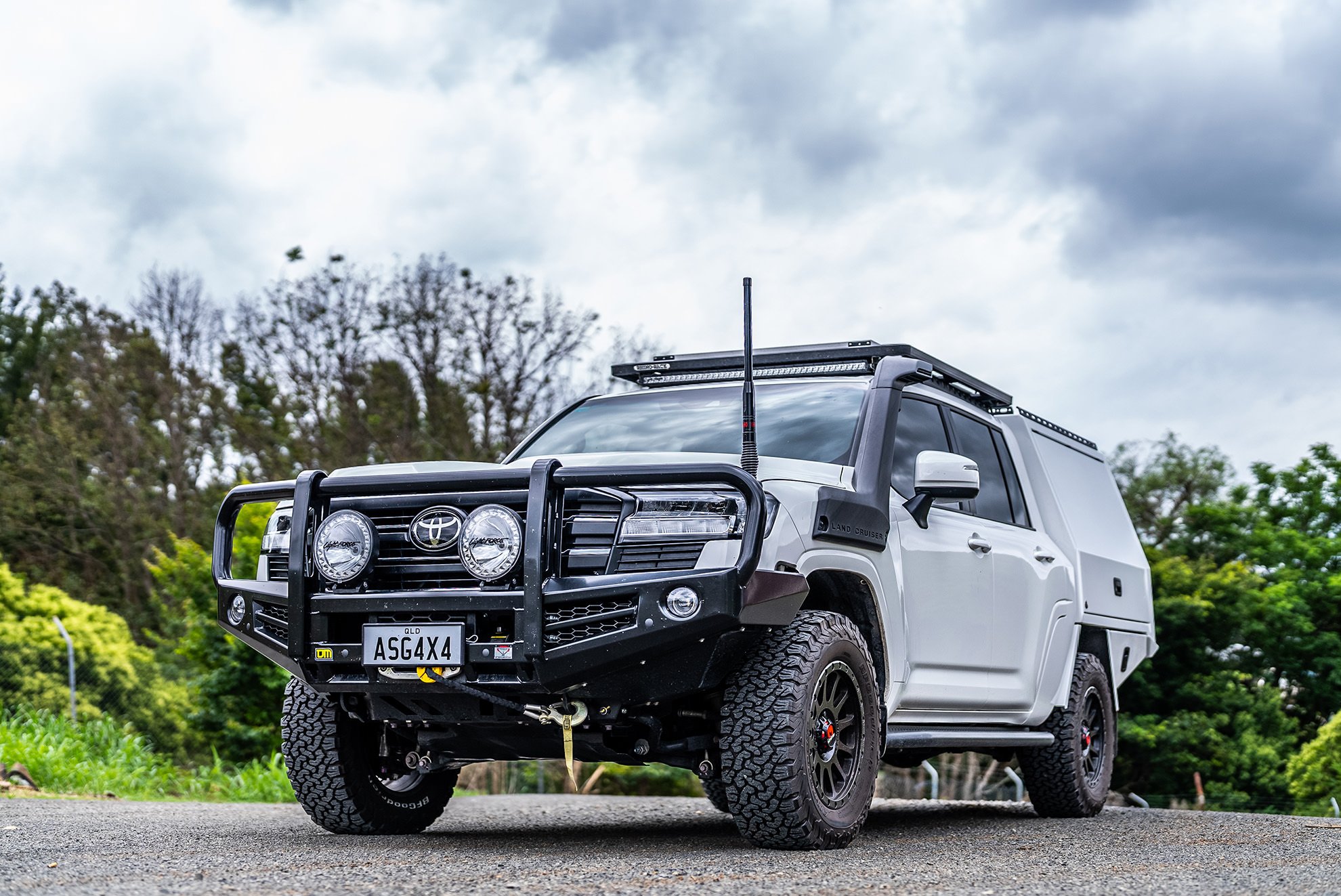 A white off-road vehicle with a black front bumper and bull bar, large tires, and roof lights, parked outdoors with green trees and cloudy sky in the background.