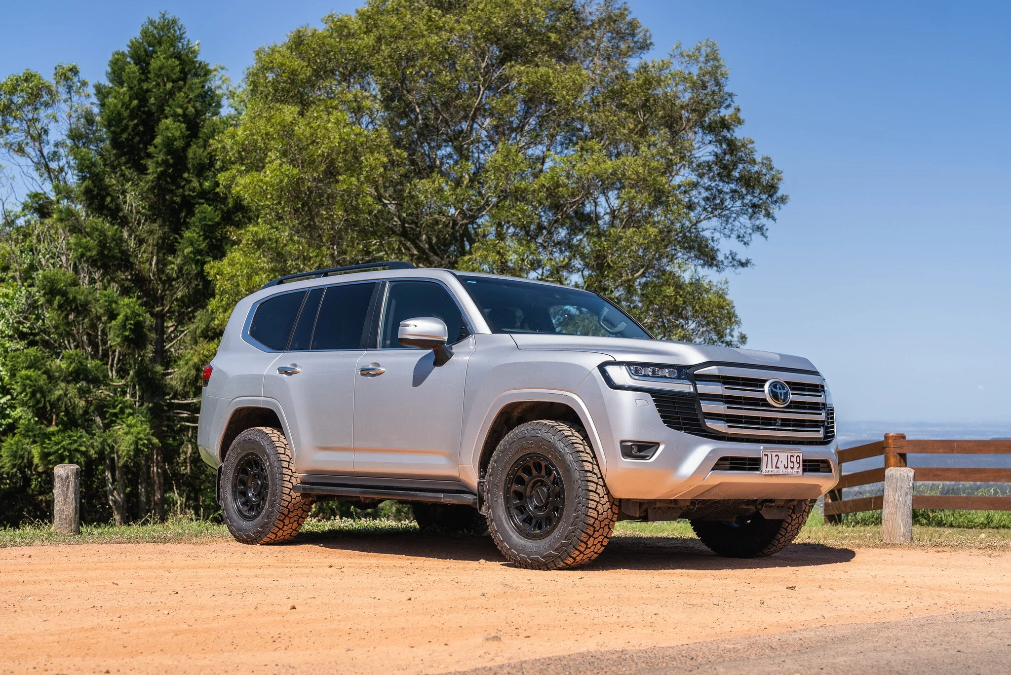 A silver Toyota SUV parked on a dirt area with green trees and a wooden fence in the background, under a clear blue sky.