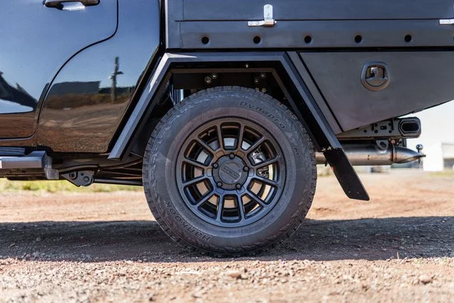 Close-up of a black off-road vehicle tire and wheel, with part of the black vehicle body visible.