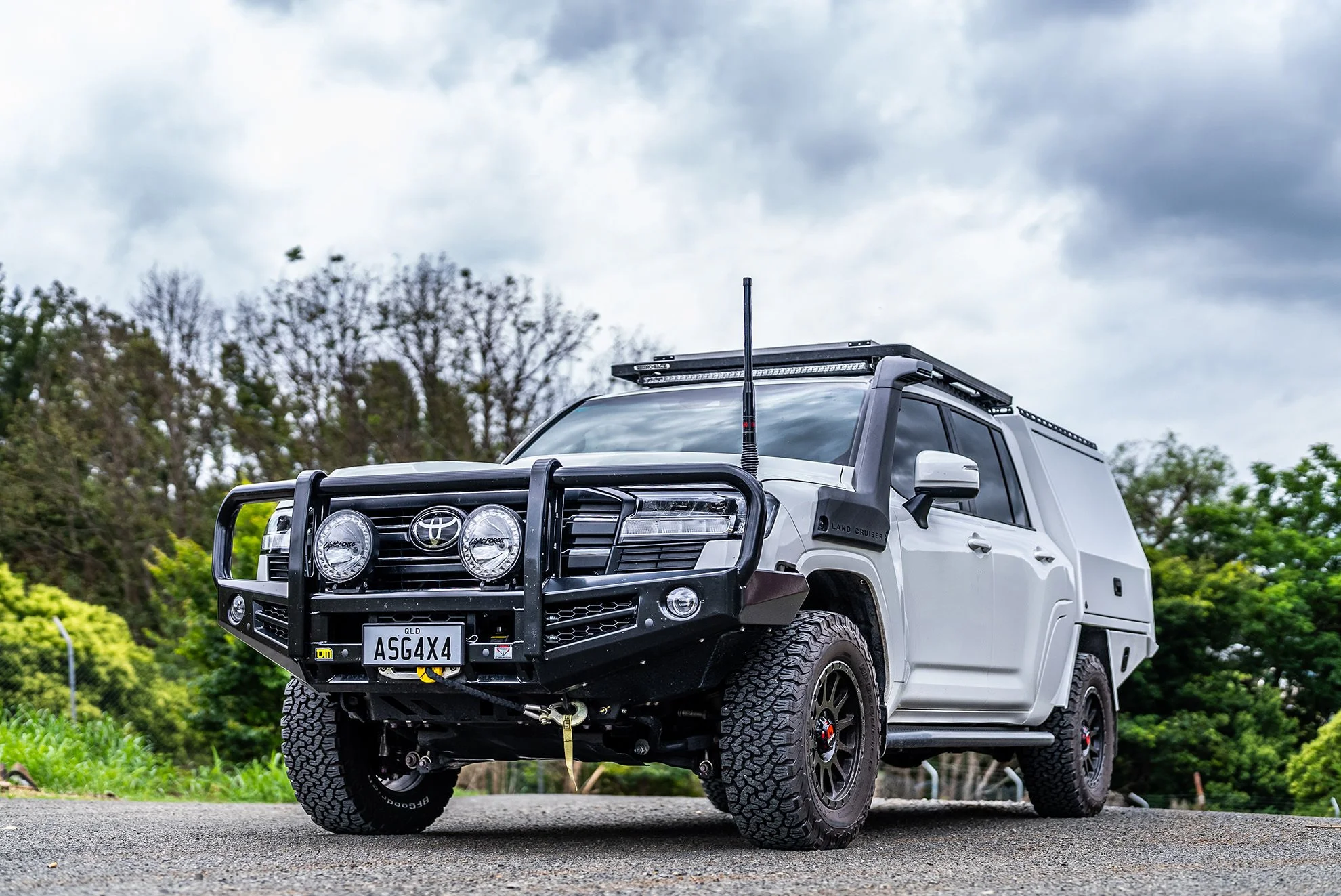 A white Toyota Land Cruiser with off-road accessories parked on a gravel road surrounded by trees and a cloudy sky.