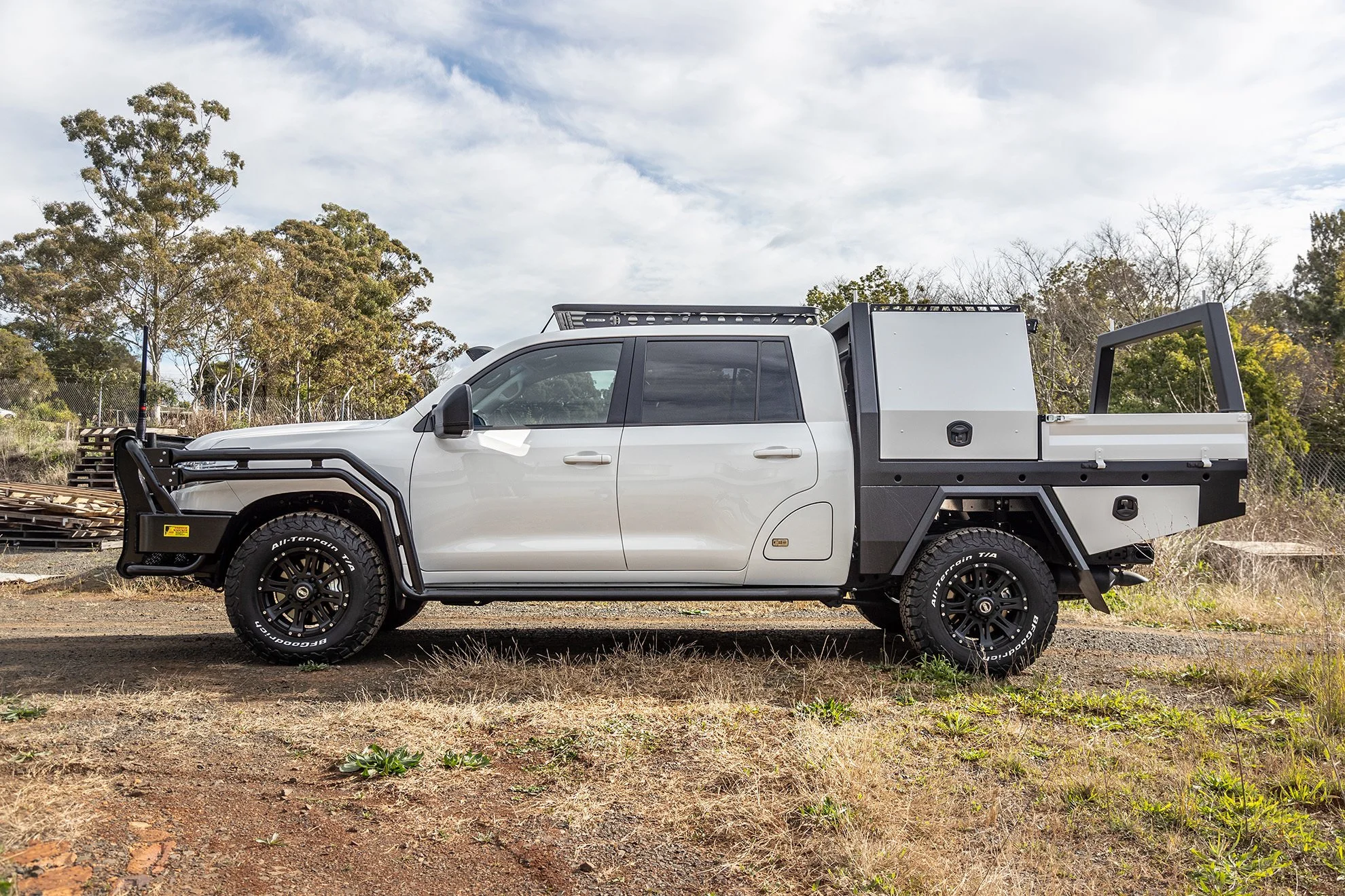 White utility truck with black off-road tires and equipment mounted in the bed, parked on a dirt lot with trees and cloudy sky in the background.