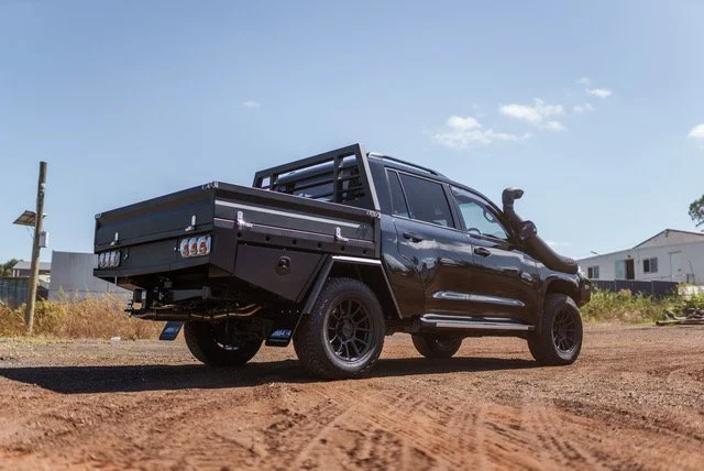 A black pickup truck with a flatbed and a roof rack parked on a dirt road with a few buildings visible in the background under a clear blue sky.