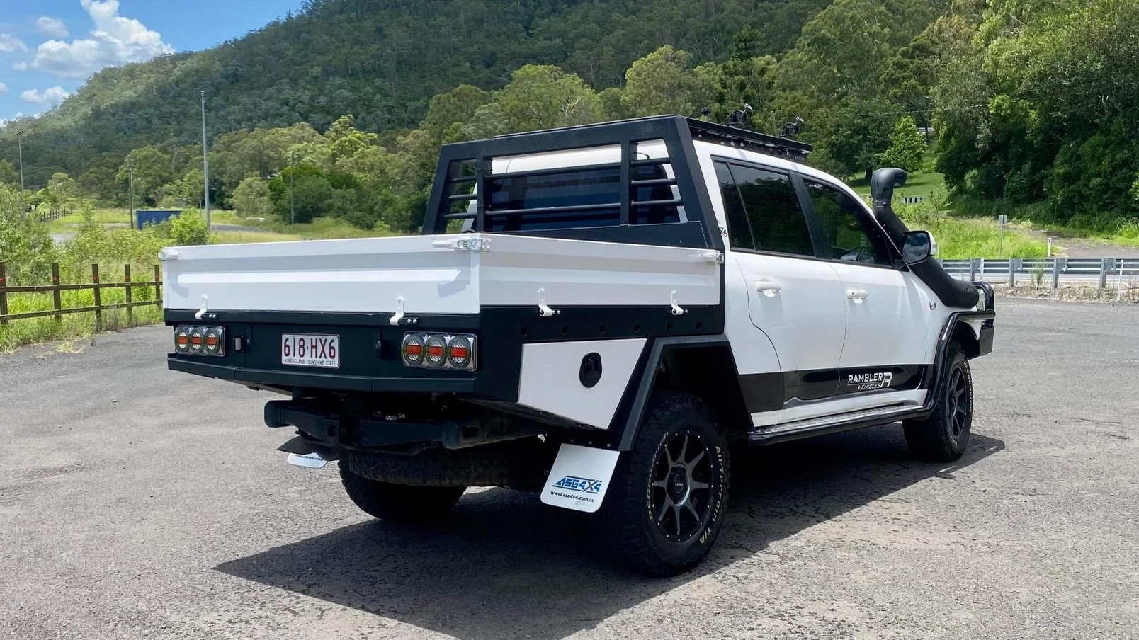 A white and black Utility Vehicle Ramble R parked on a gravel surface, with a black canopy and a power snorkel on the side, behind the cab, in a rural setting with green trees and hills in the background.
