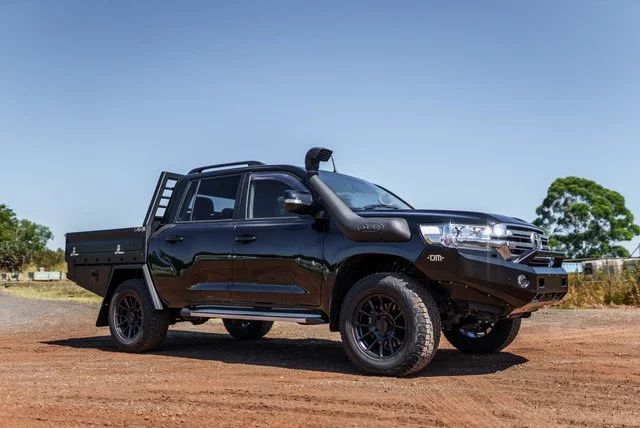 Black pickup truck with off-road tires parked on dirt ground under clear blue sky, with trees in the background.