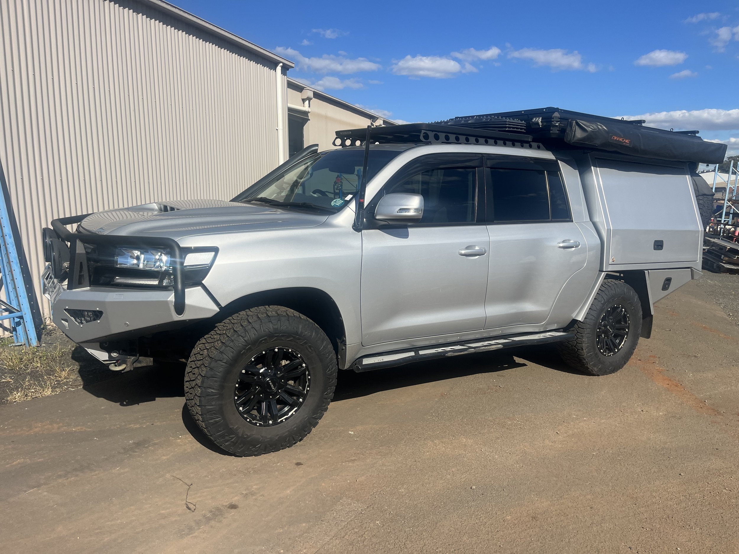 Silver truck with off-road tires, a large roof rack, and a custom camper shell on the back, parked on a dirt lot next to a beige metal building under a partly cloudy sky.