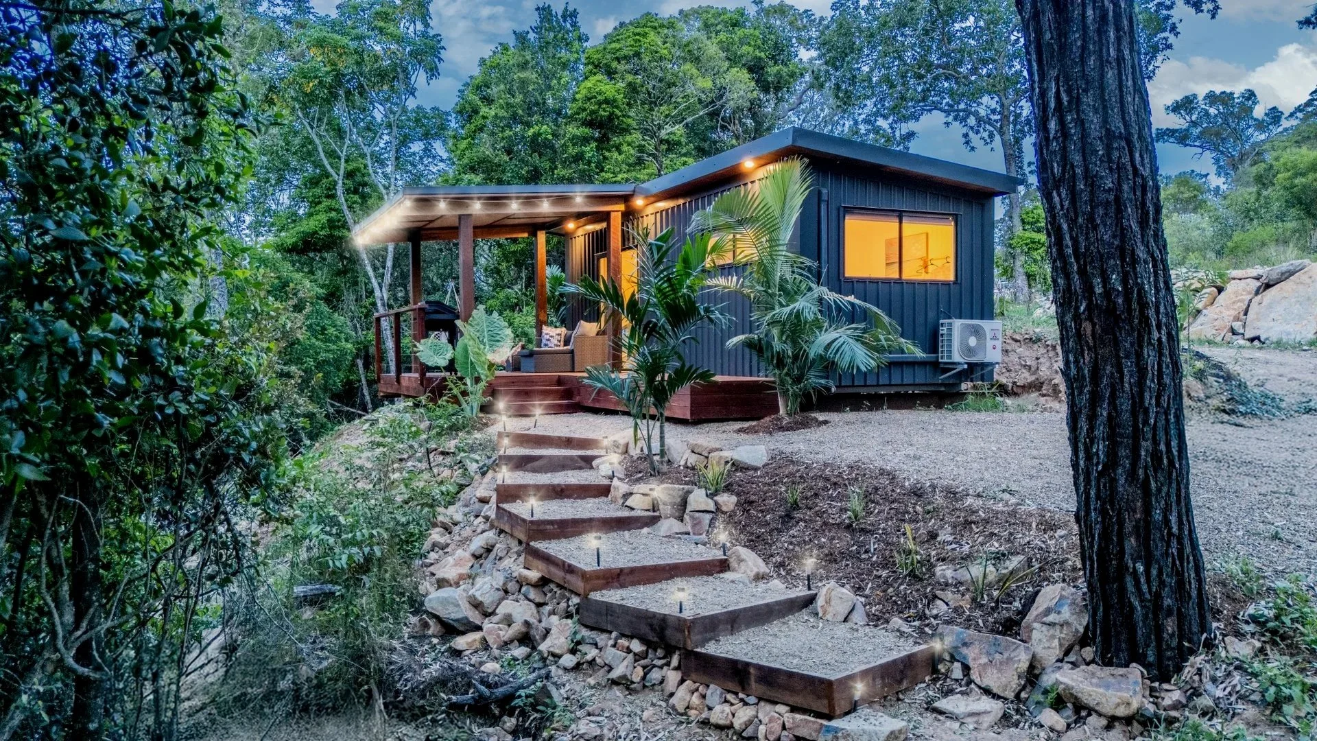 A modern tiny house with a dark exterior, large windows, and a wooden porch in a wooded area, with a stone and wooden staircase leading to the entrance, surrounded by greenery and trees.