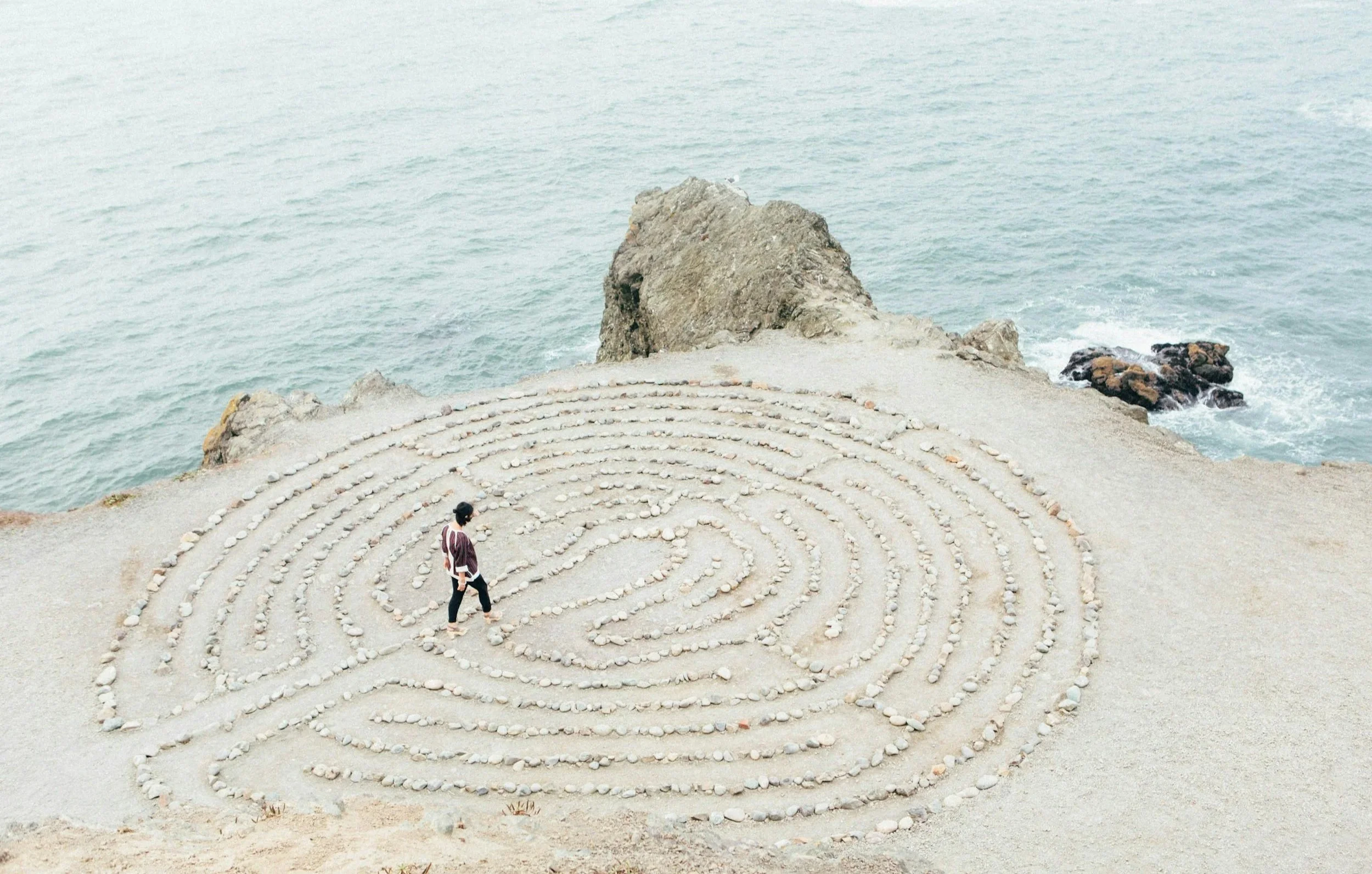 A person walking through a spiral of rocks (a labyrinth) on a sandy beach near the ocean, with a rocky outcrop in the background.