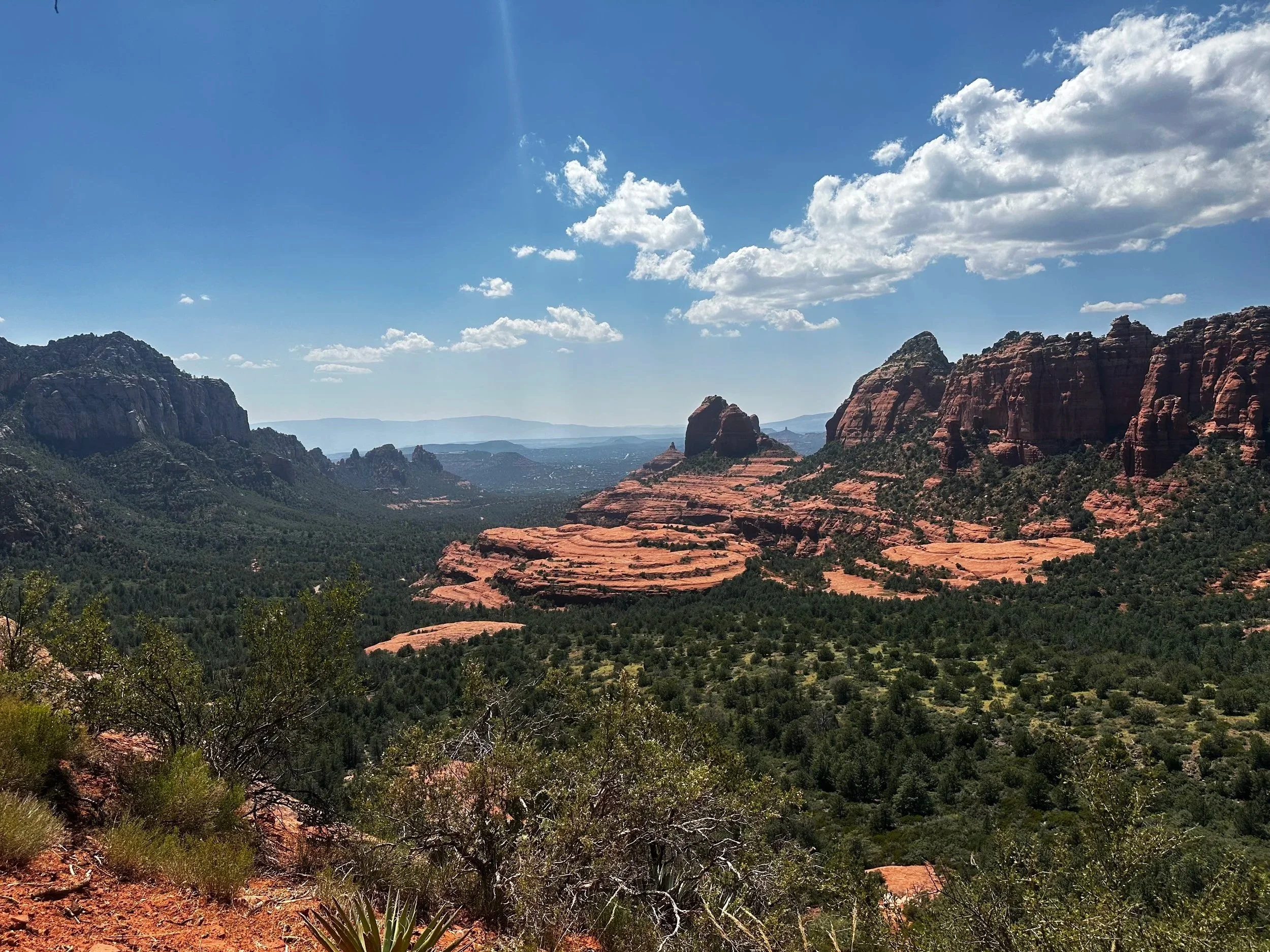 A panoramic view of Sedona's red rock formations under a blue sky with scattered clouds, lush green vegetation in the foreground.