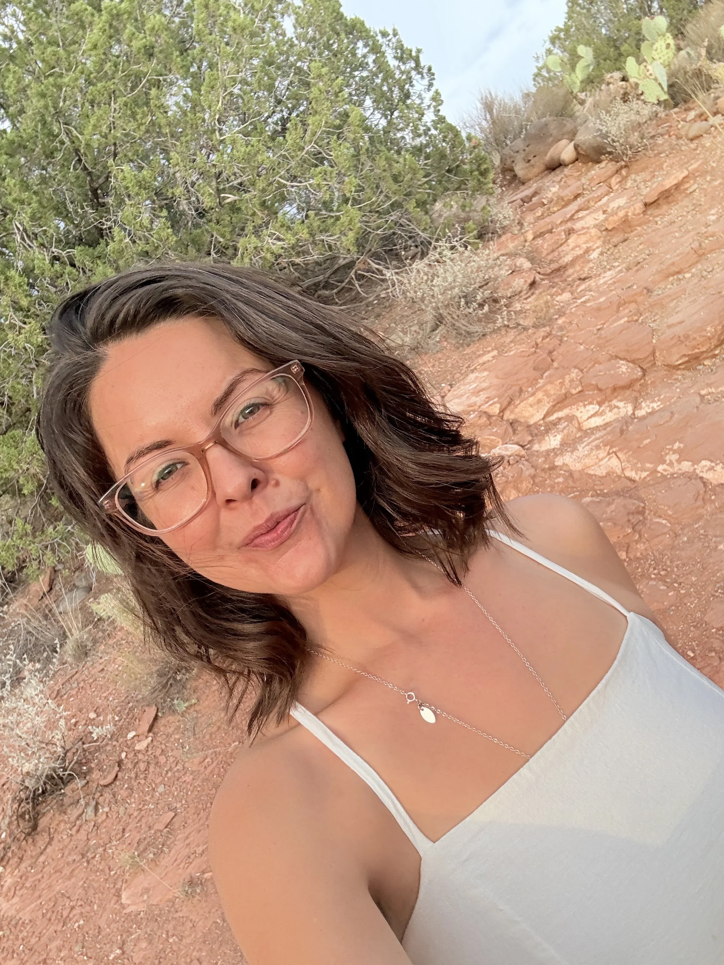 A woman with glasses and shoulder-length brown hair taking a selfie outdoors against a desert landscape with red rocks, green trees, and cacti.