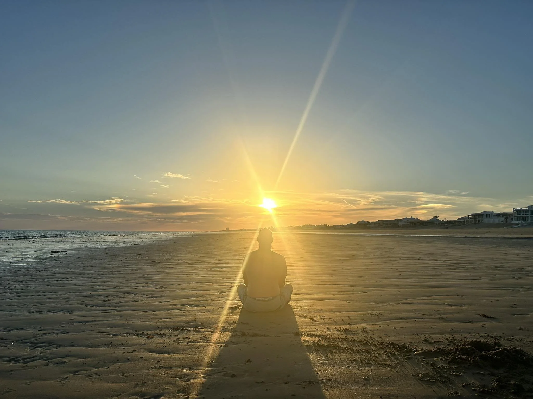 A person sitting cross-legged on the beach, facing the sunset, with the sun just above the horizon and a clear sky with a few clouds.