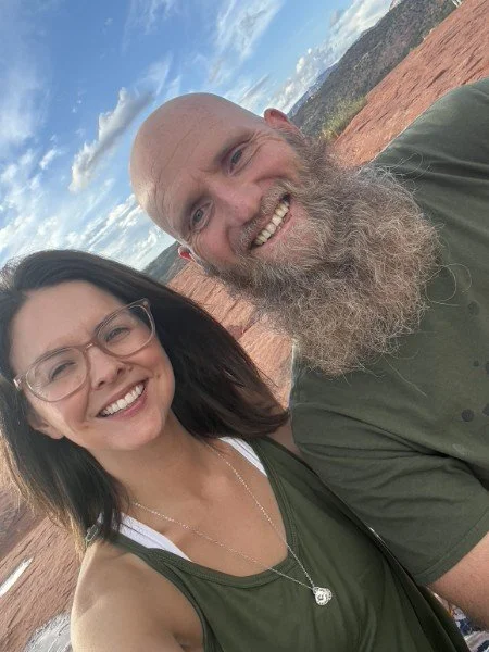 A smiling woman with glasses and a beaded necklace taking a selfie with a smiling bald man with a long beard, outdoors with a red rock landscape and blue sky with clouds in the background.