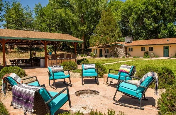 Outdoor patio with six blue cushioned chairs surrounding a fire pit, a pavilion with a barbecue area, and a small building in the background with trees and greenery.