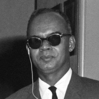 Black and white photograph of a man wearing sunglasses, a suit, and a tie, with headphones in his ears.