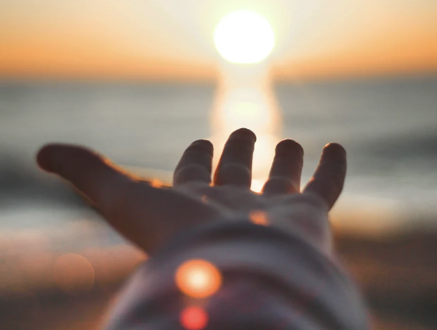 A hand reaching out toward a sunset over the ocean, with the sun's reflection on the water.