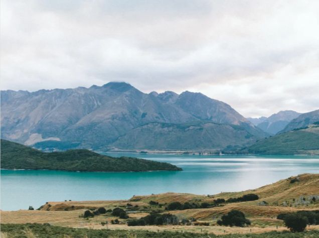 Scenic view of a lake surrounded by mountainous terrain, with grassy hills in the foreground and mountains in the background under a cloudy sky.
