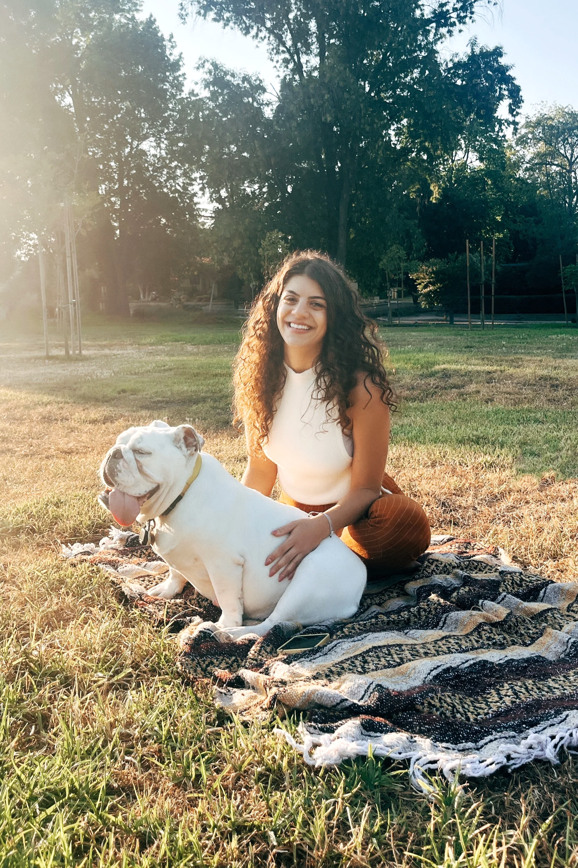 A young woman with curly hair smiling and sitting on a blanket in a park with a happy bulldog