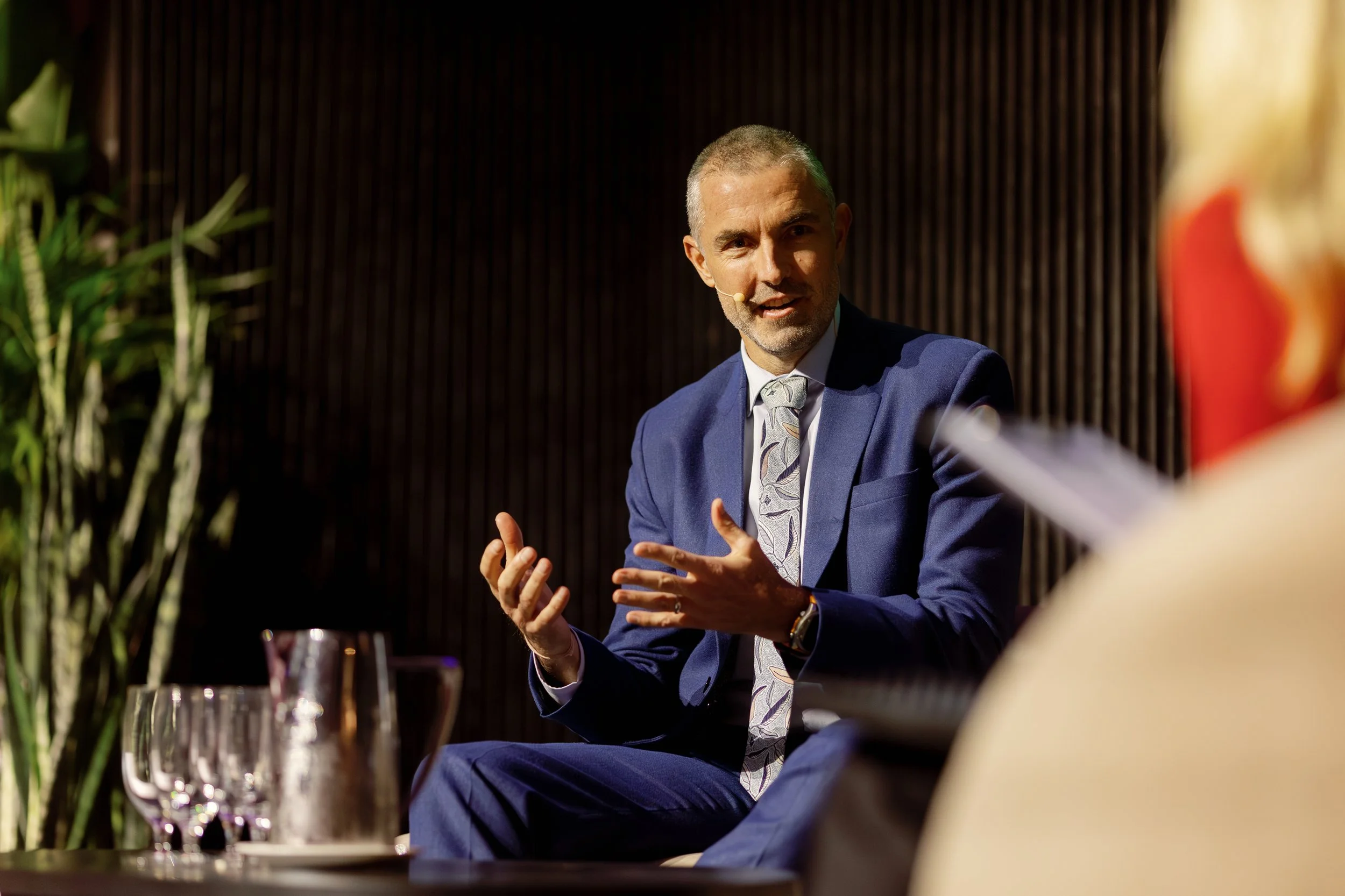 Freight Forward A man in a blue suit, white shirt, and patterned tie speaking at an event with a microphone headset, gesturing with his hands, seated at a table with wine glasses and a plant in the background.
