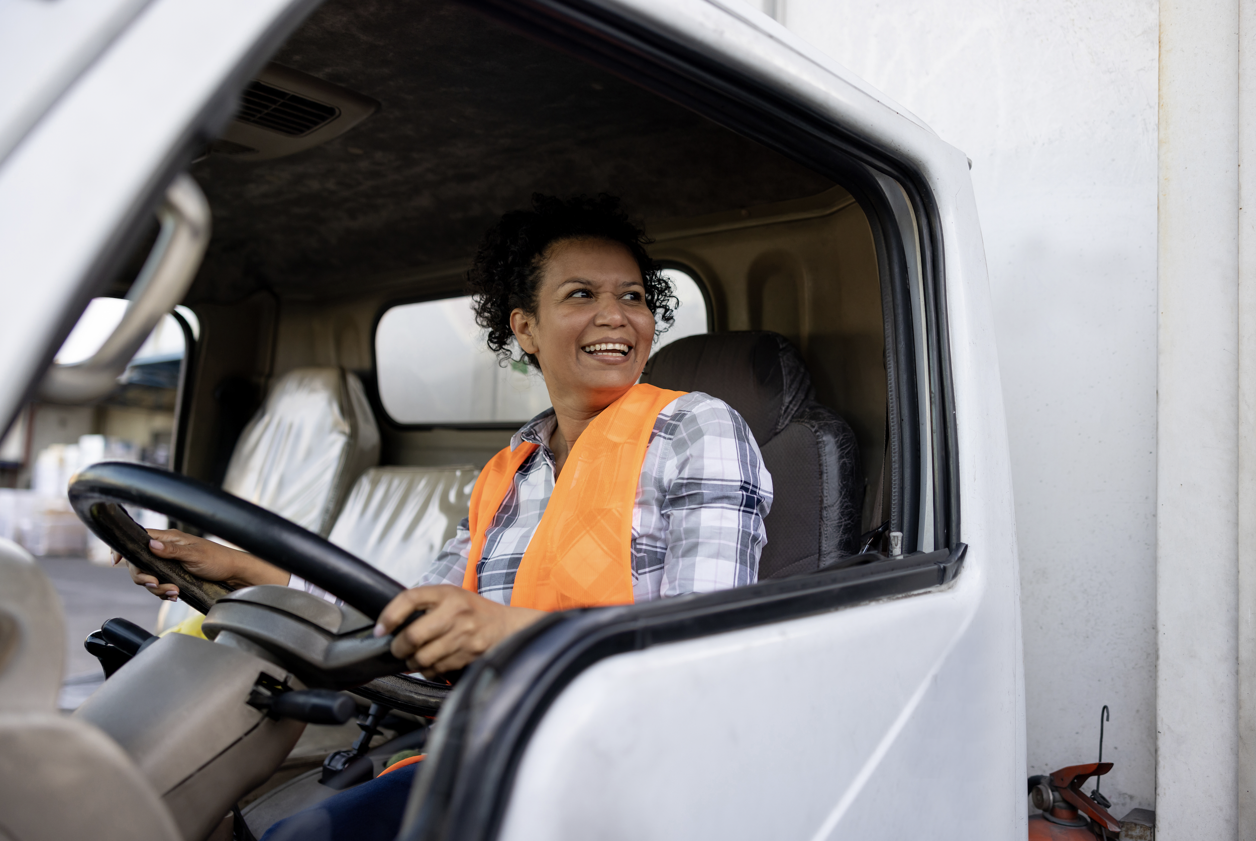 Woman sitting in the driver's seat of a truck, smiling, wearing an orange safety vest and plaid shirt, with the truck's interior dashboard visible.