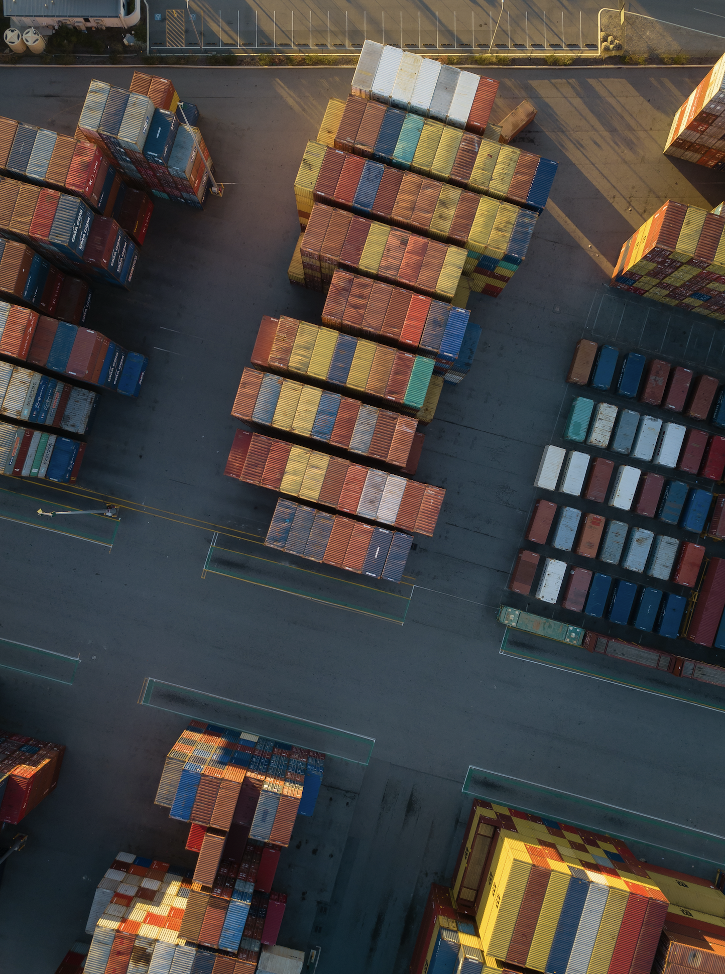 Aerial view of stacked shipping containers in a port, with some parked vehicles and parking spaces visible.