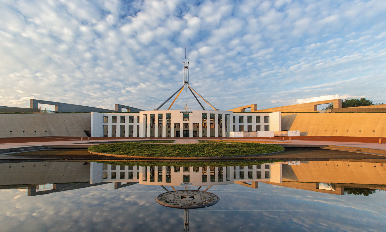 The Australian Parliament House with a reflecting pool in front, under a partly cloudy sky.
