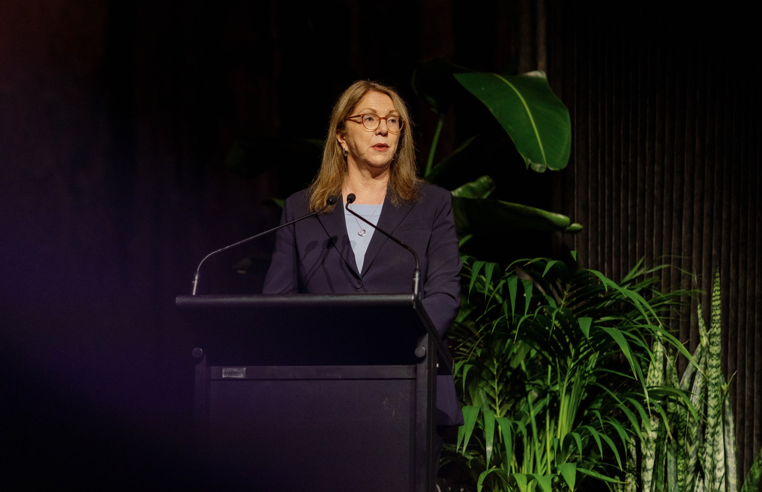 A woman wearing glasses and a dark blazer is standing at a podium with two microphones, giving a speech or presentation. There are green plants in the background.
