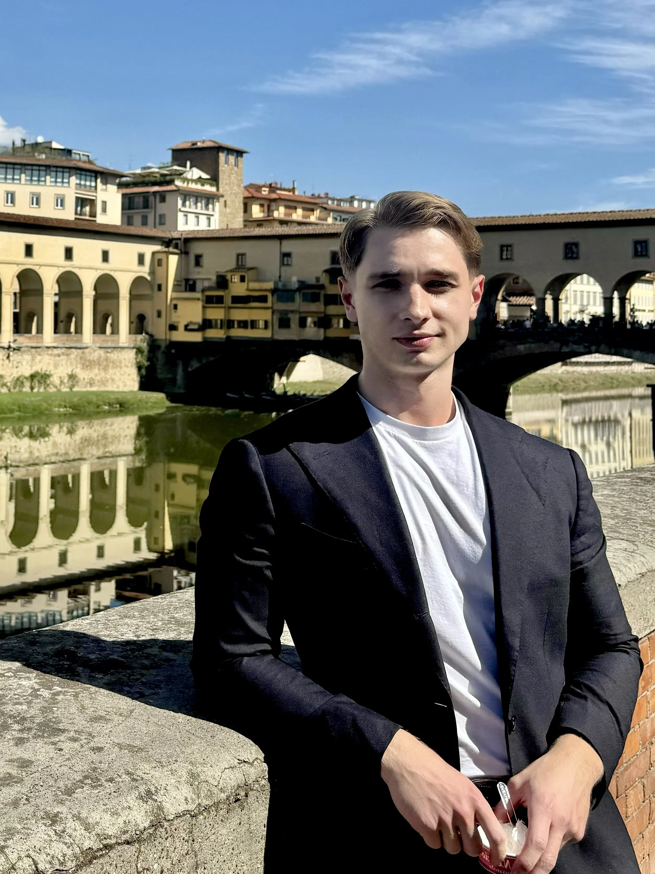 A portrait of Luke Kosonen in a black blazer and white t-shirt standing outdoors with a cityscape on a sunny day.
