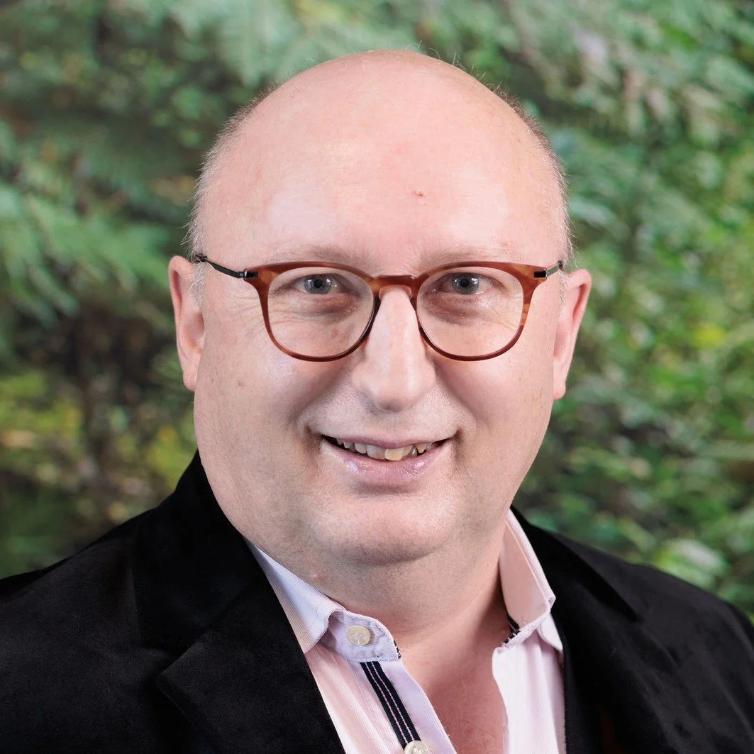 Headshot of a bald man with glasses, wearing a black jacket and a button shirt, smiling against a blurred plant green background.