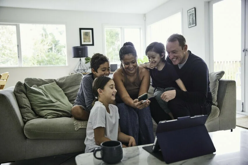 A family of five sitting together on a sofa in a brightly lit living room, looking at a smartphone held by the mother. There is a tablet and a mug on the coffee table in the foreground.