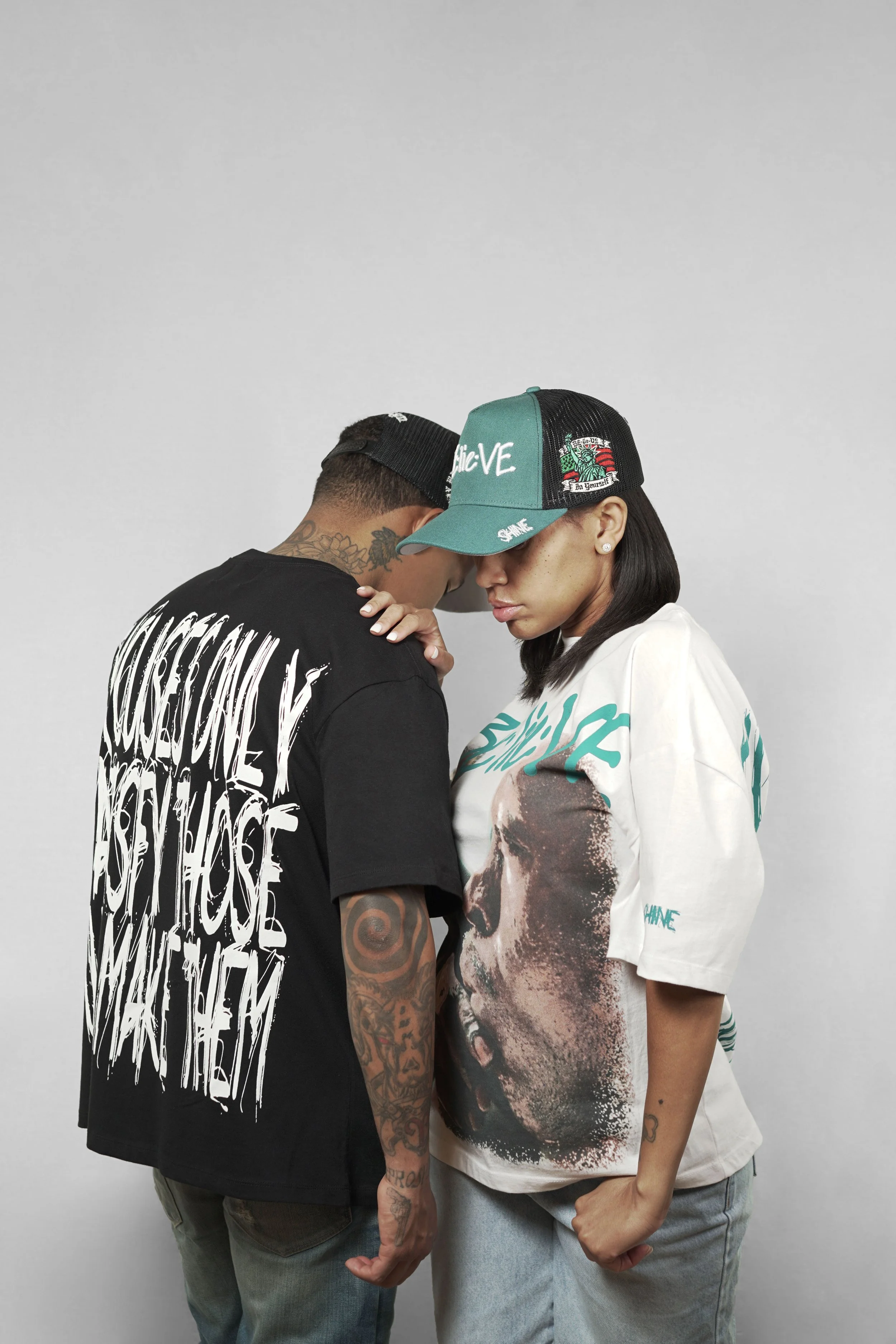 Two people face each other closely, their foreheads touching, with one person's hand on the other's shoulder. They wear casual clothing and caps, with a plain gray background.