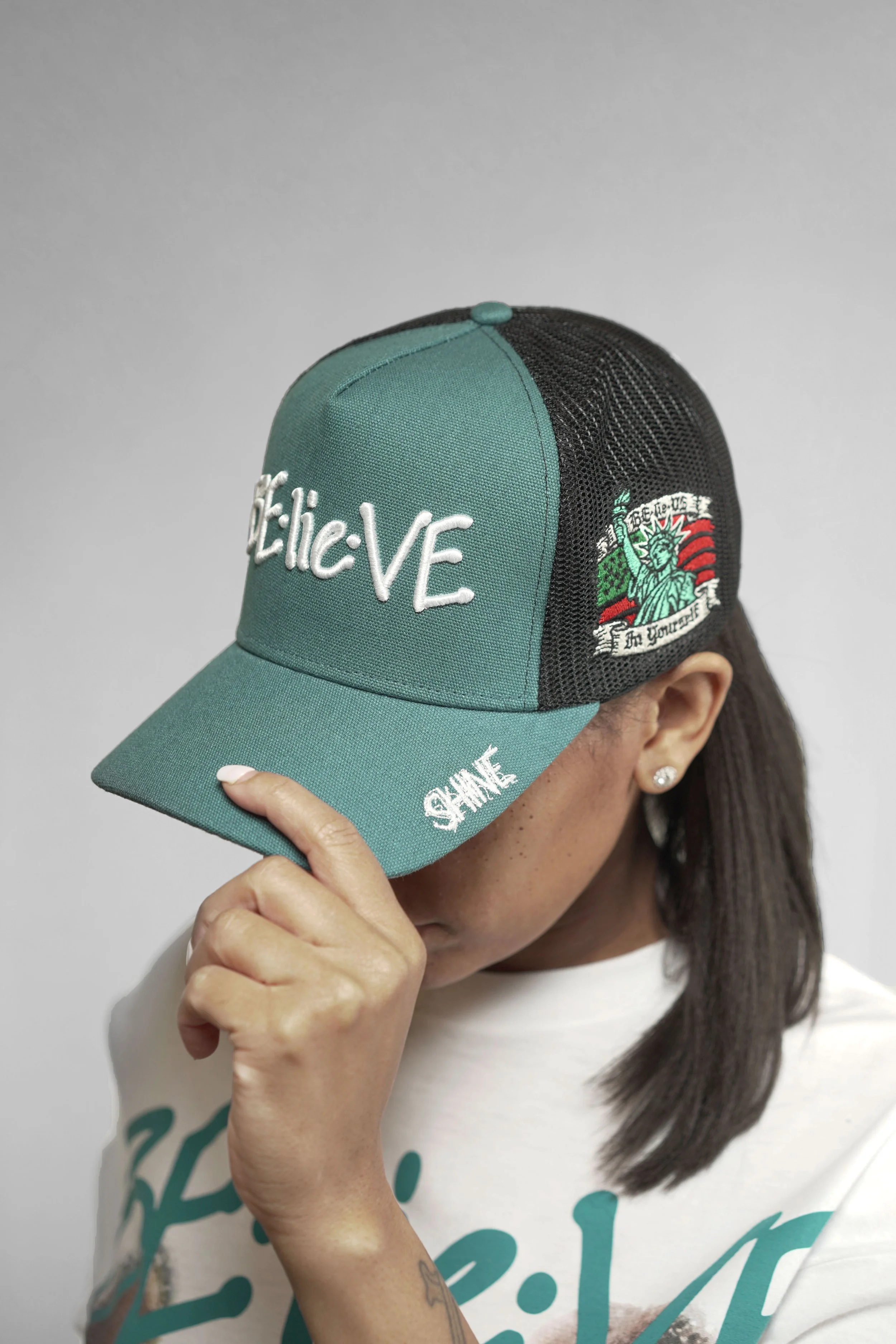 Person wearing a baseball cap with embroidered text and a patch, holding the brim with fingers, showing part of a white T-shirt with teal lettering, long dark hair, earrings, and a tattoo on the arm, standing against a plain gray background.