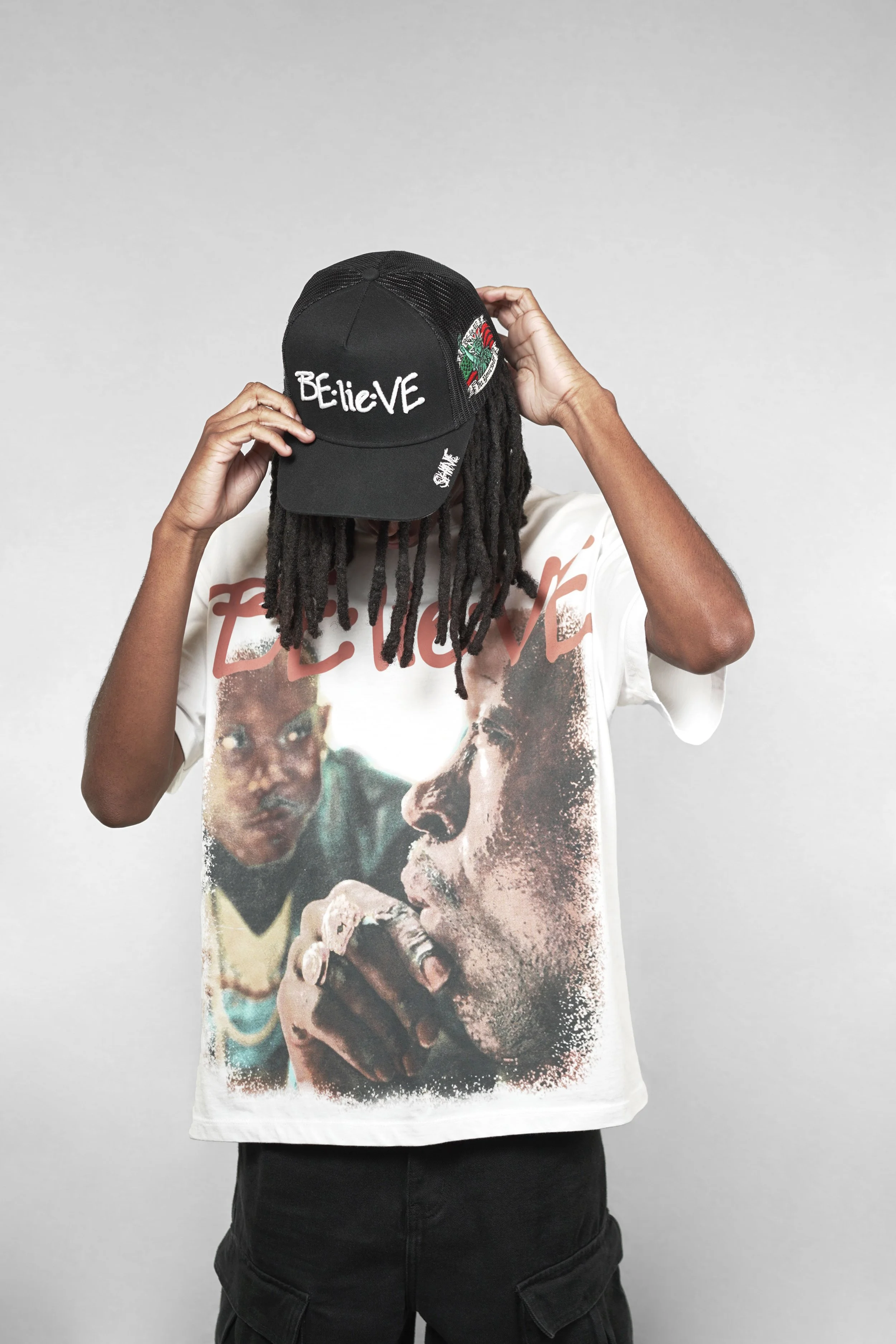 Person wearing a black cap with "Believe" embroidered on the front, a graphic T-shirt featuring a portrait of a woman smoking, and black cargo pants, standing against a plain gray background.