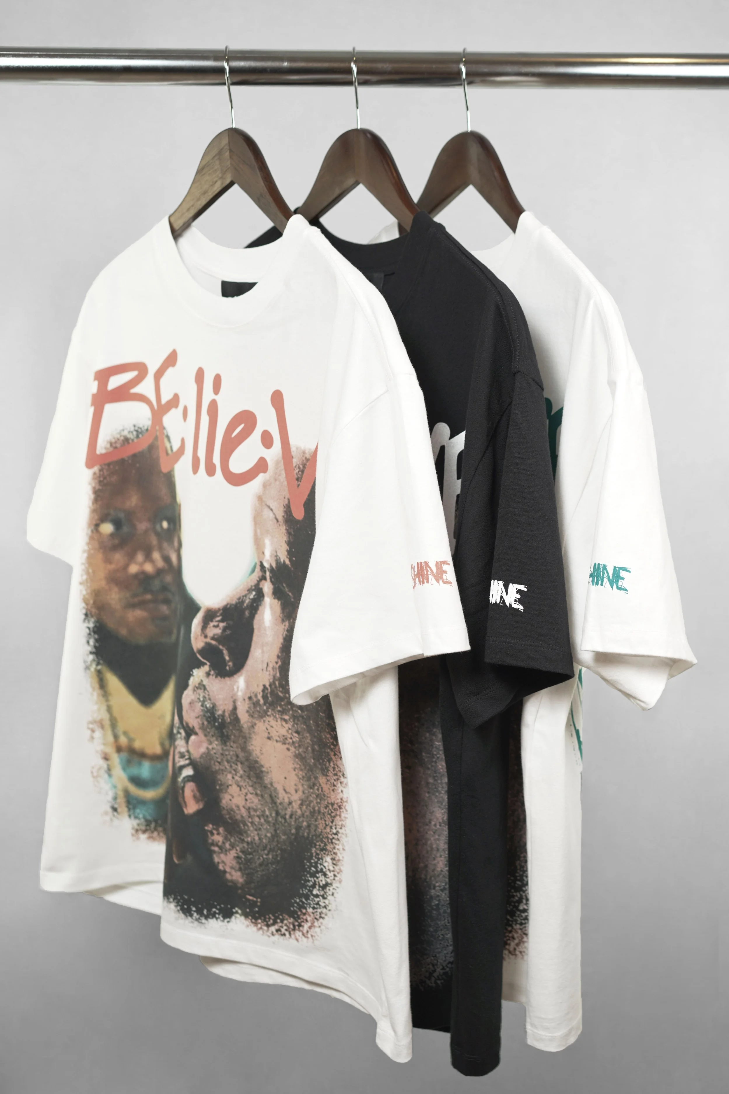 Three T-shirts hanging on wooden hangers on a metallic clothing rack. The first T-shirt is white with a colorful graphic at the front. The second T-shirt is black, and the third T-shirt is white with colorful text on the sleeve.